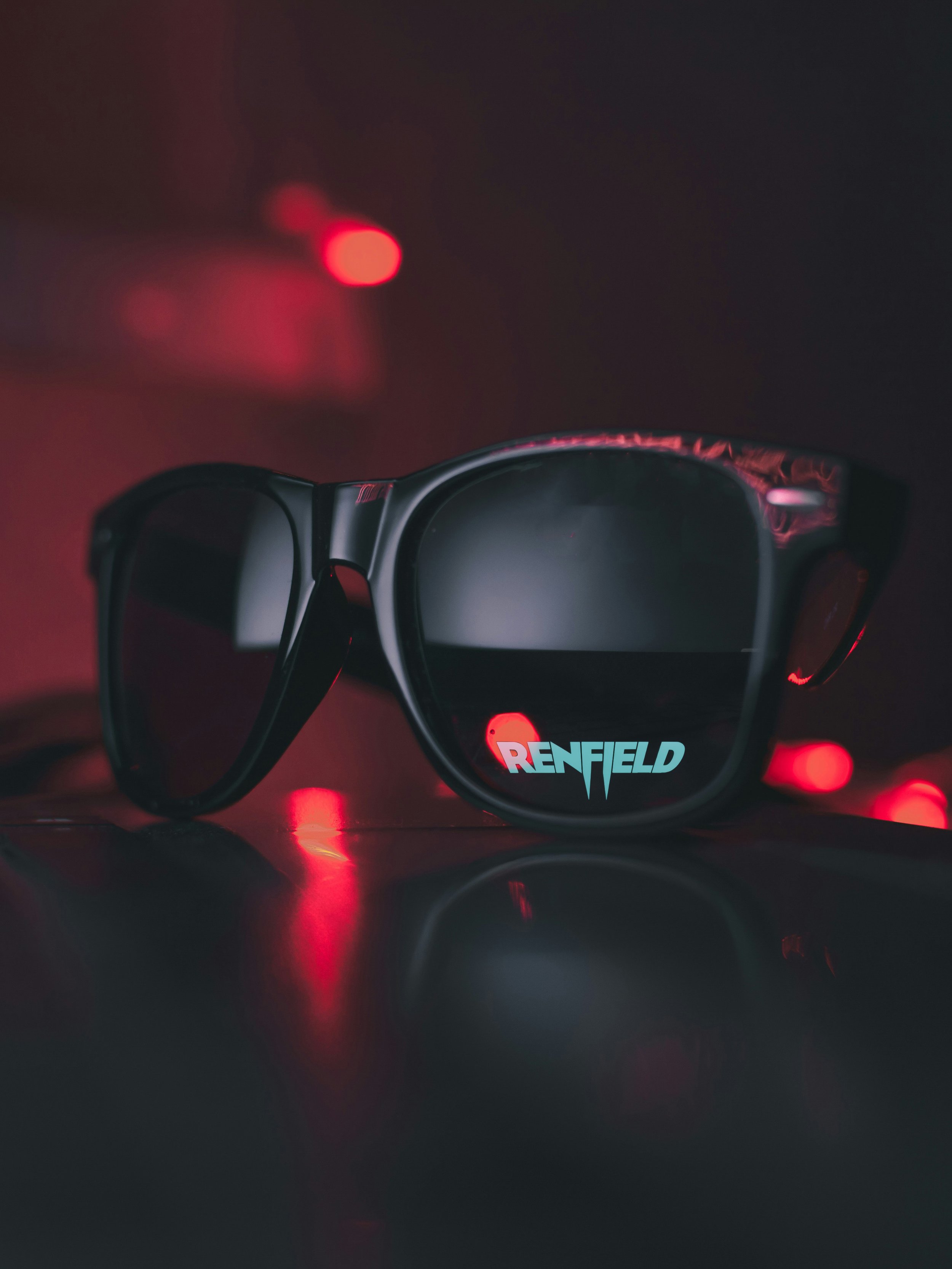 A pair of black sunglasses resting on a dark surface, with red reflections and a blurred red light in the background. The reflection in the glasses shows the text 'RENFIELD' in light blue and red.