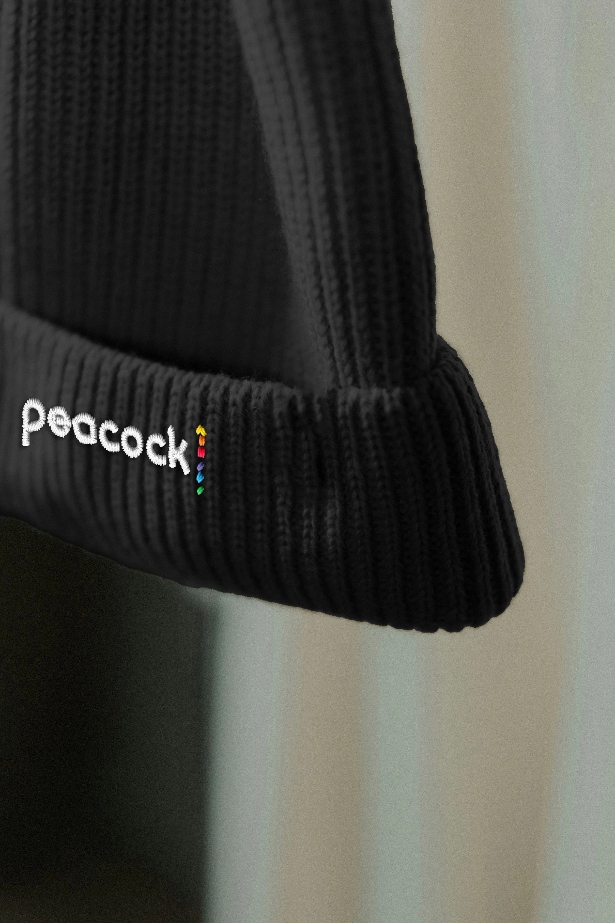 Close-up of a black, ribbed knit beanie with the brand name 'pocock' embroidered in white and highlighted with rainbow-colored dots.