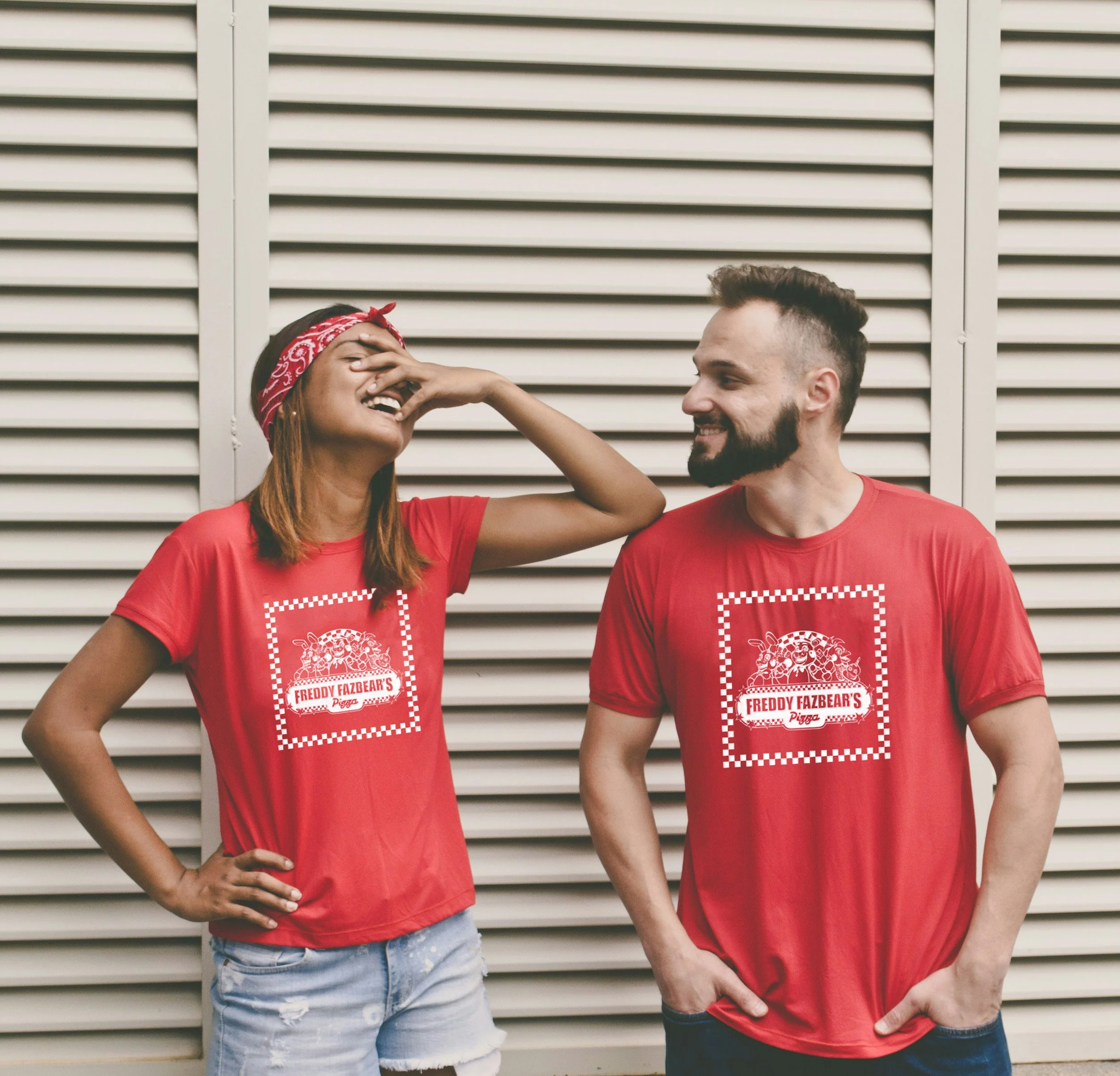 Two people wearing red T-shirts with a logo, standing in front of a metal louvered wall. One person, a woman, is laughing and covering her face with her hand, while the other, a man, is smiling and looking at her.