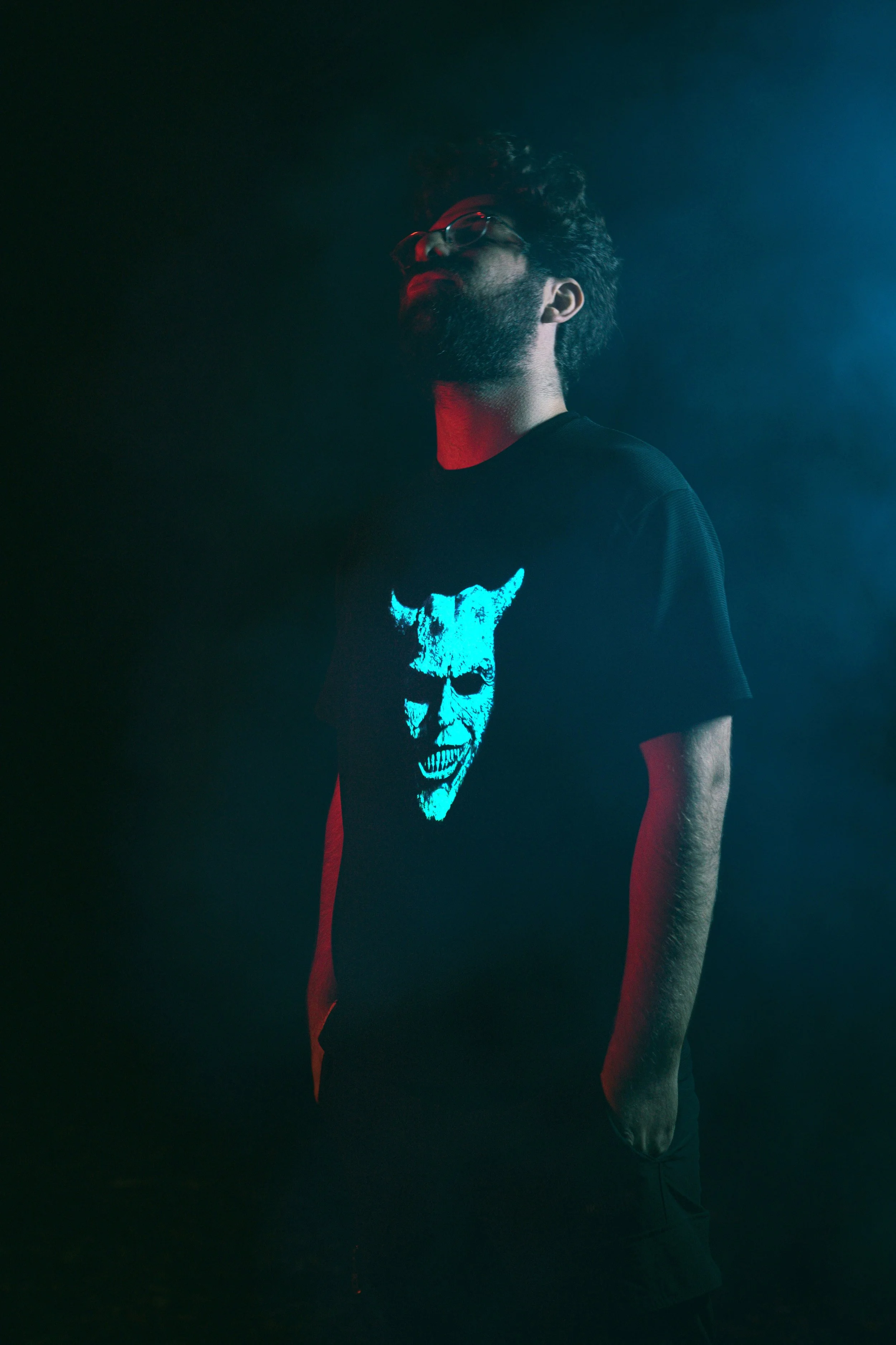 A man with glasses and a beard wearing a black T-shirt with a glowing blue demon face design, standing in a dark, smoky environment with red and blue lighting.