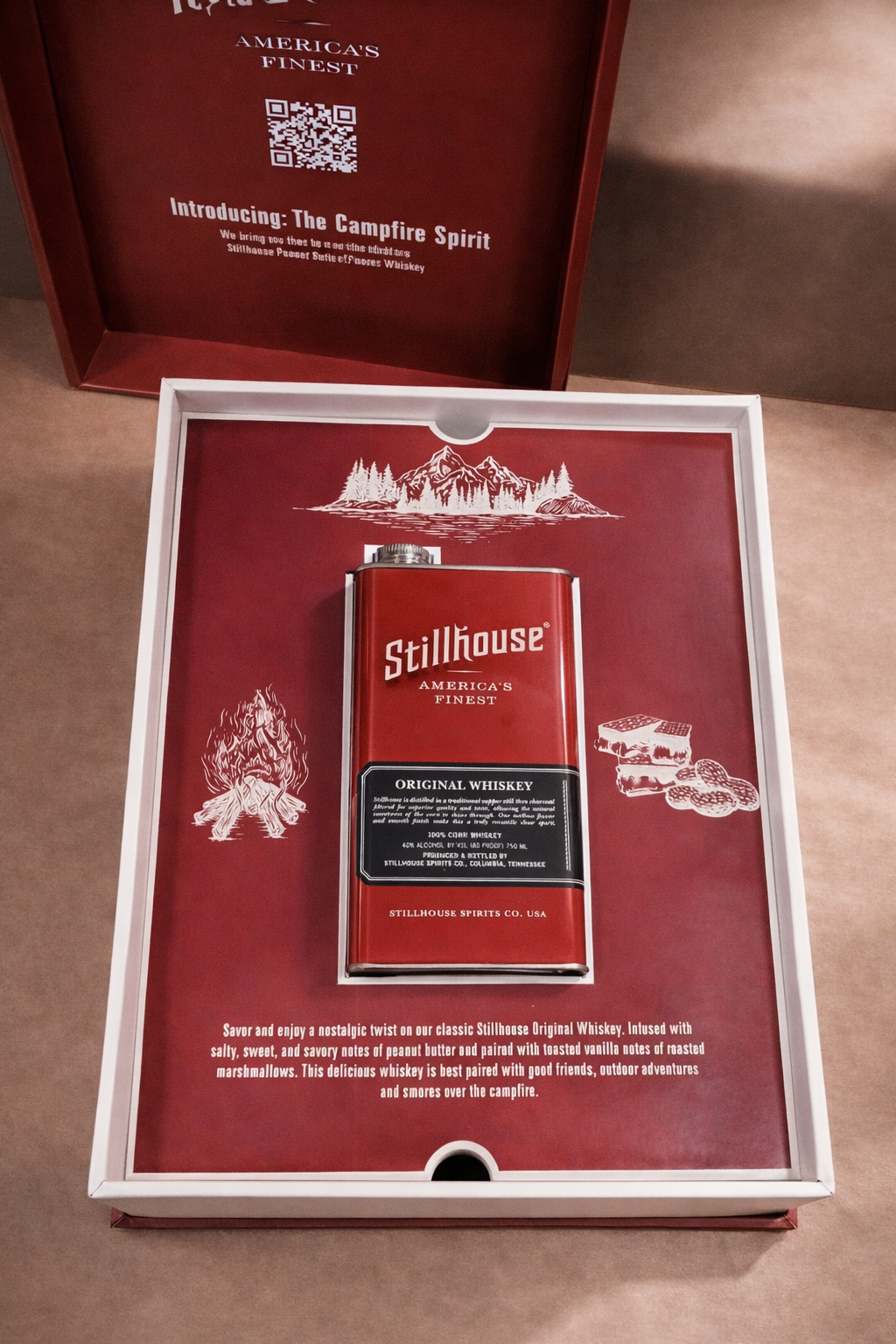 A red box with Stillhouse America's Finest whiskey bottle in a see-through window, surrounded by illustrated mountain, trees, campfire with logs, and cookies, with text describing the whiskey's flavor and origin inside the box.