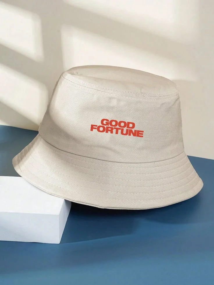 A beige bucket hat with the words "GOOD FORTUNE" printed in red on the front, placed on a blue surface next to a white rectangular object.