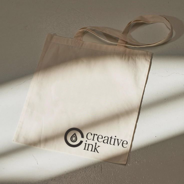 A beige tote bag with the words "Creative Ink" and a logo printed in black, placed on a white surface with a shadow cast across it.