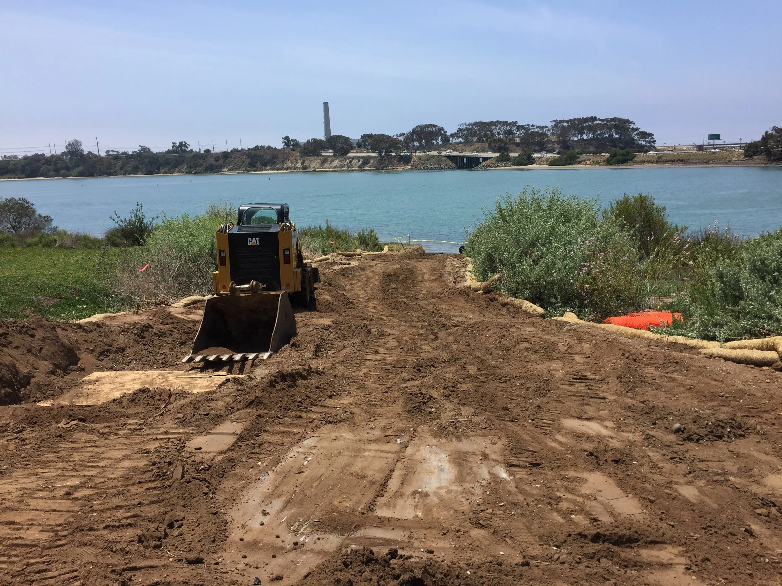 Construction site near a body of water with a small yellow bulldozer and cleared dirt path, bushes on sides, blue water and distant shoreline in background.
