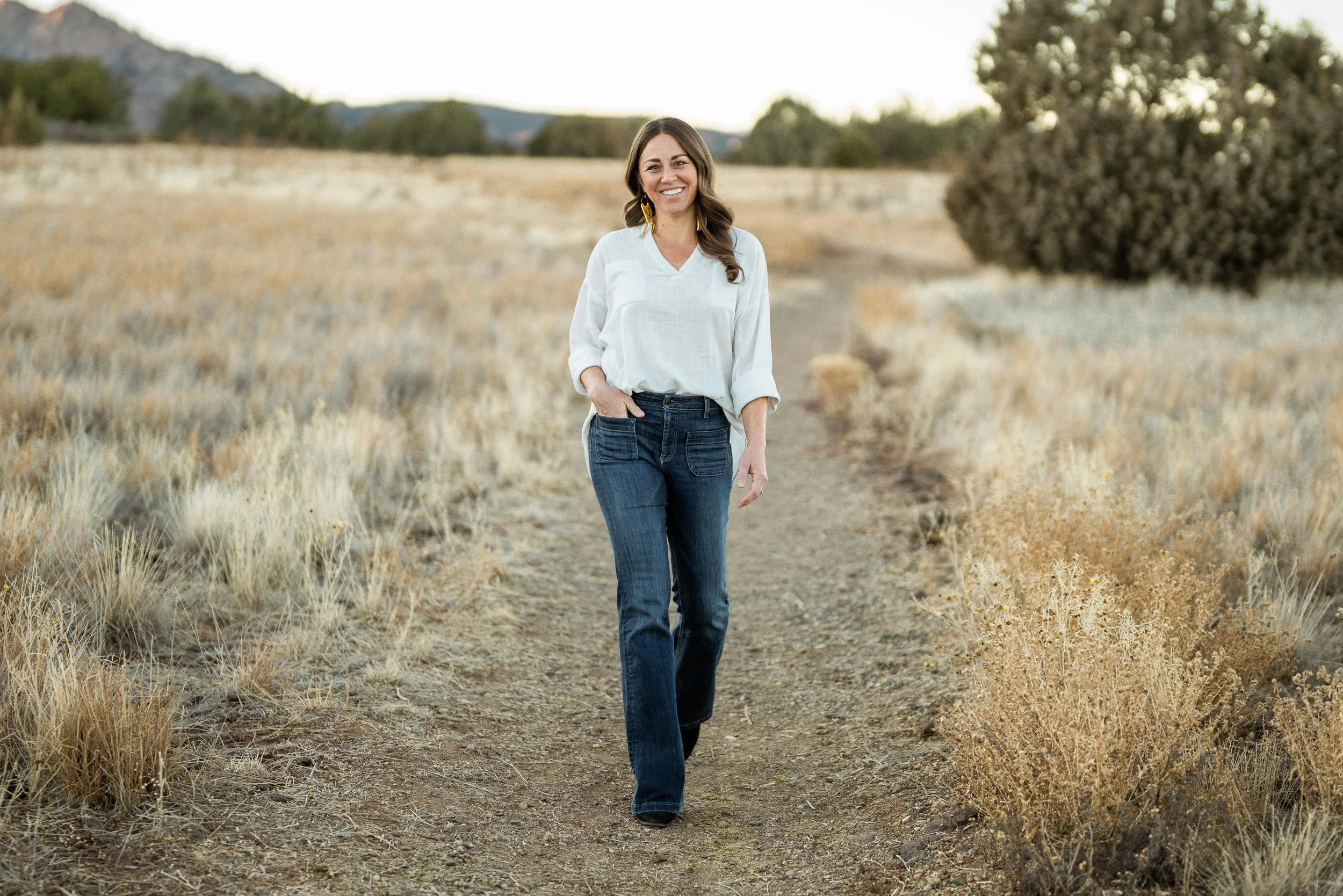 A woman with long brown hair, dressed in a white blouse and blue jeans, walking on a dirt path through a dry, grassy landscape with trees and hills in the background, smiling at the camera.