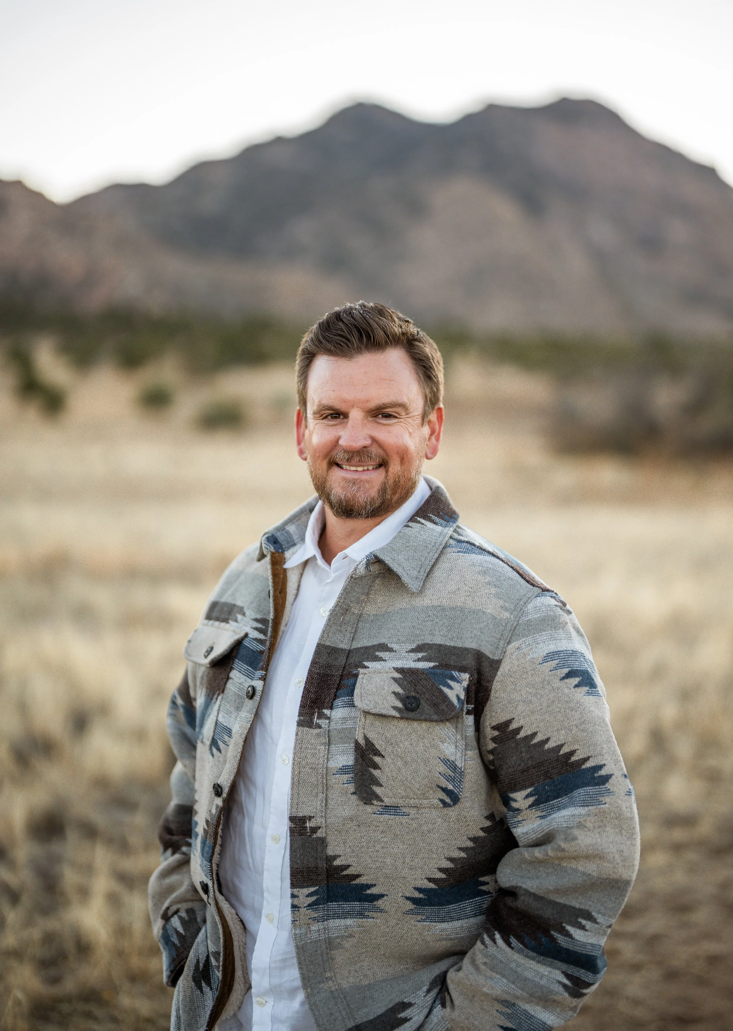 A smiling man in a patterned jacket standing outdoors in a desert landscape with mountains in the background.