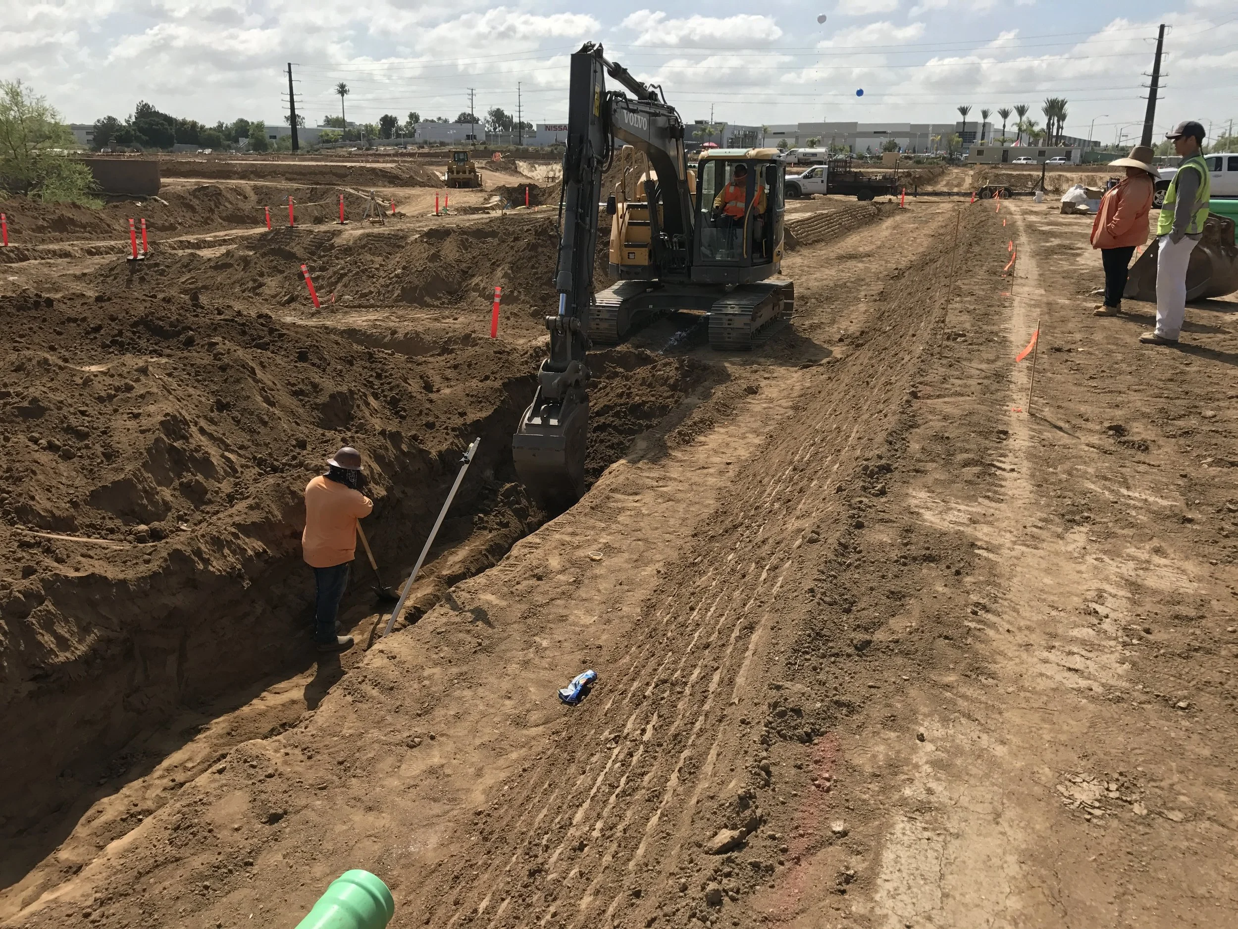 Construction site with workers and heavy machinery working on excavation, with a person using a shovel, orange markers, and other workers observing.