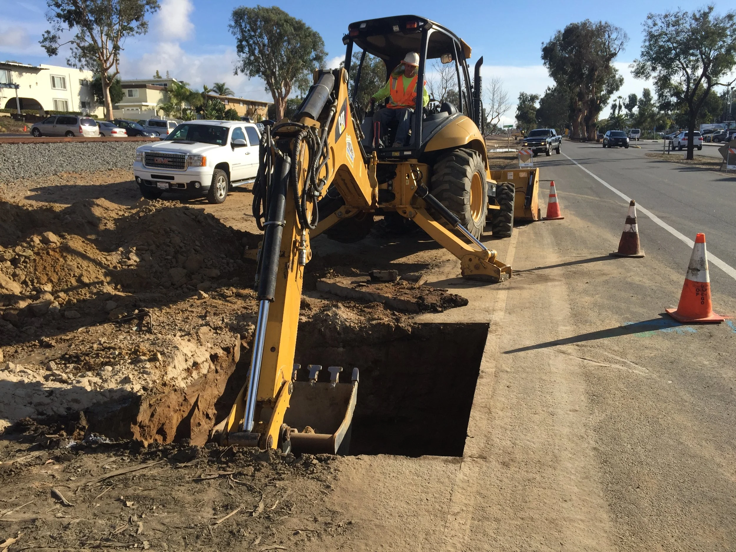 Construction worker operating a backhoe excavator digging a trench along a city street, surrounded by traffic cones.
