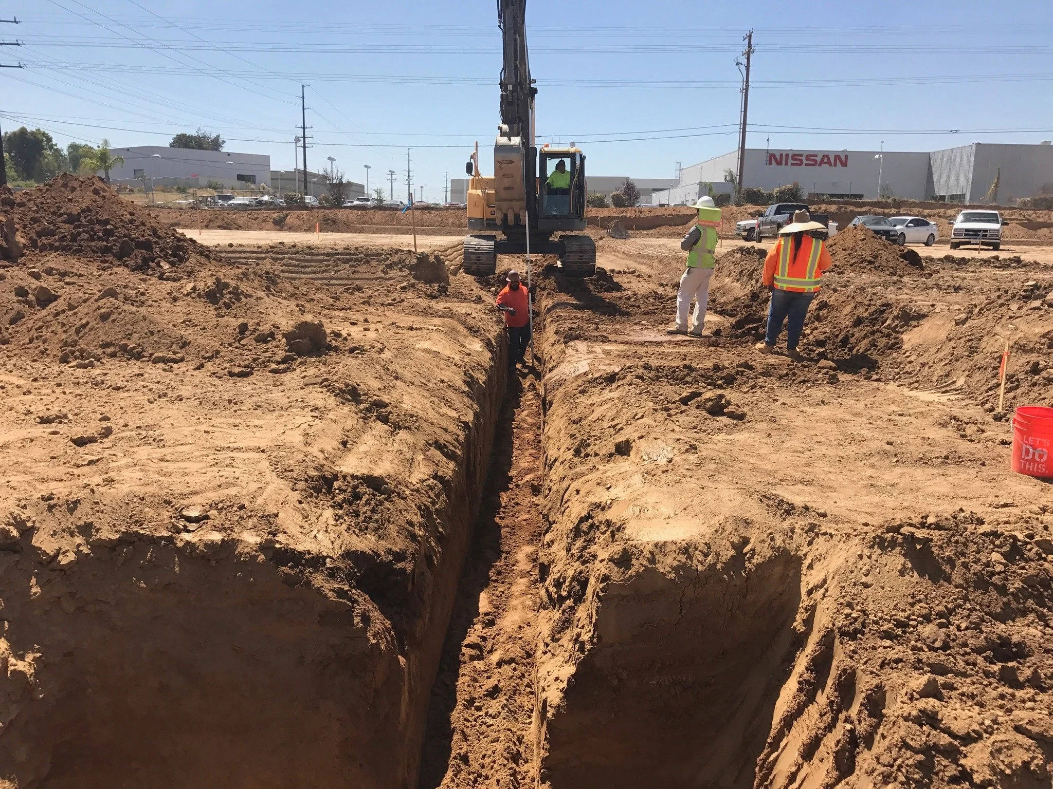 Construction workers and an excavator working on a large trench at a construction site, with several cars and buildings visible in the background.