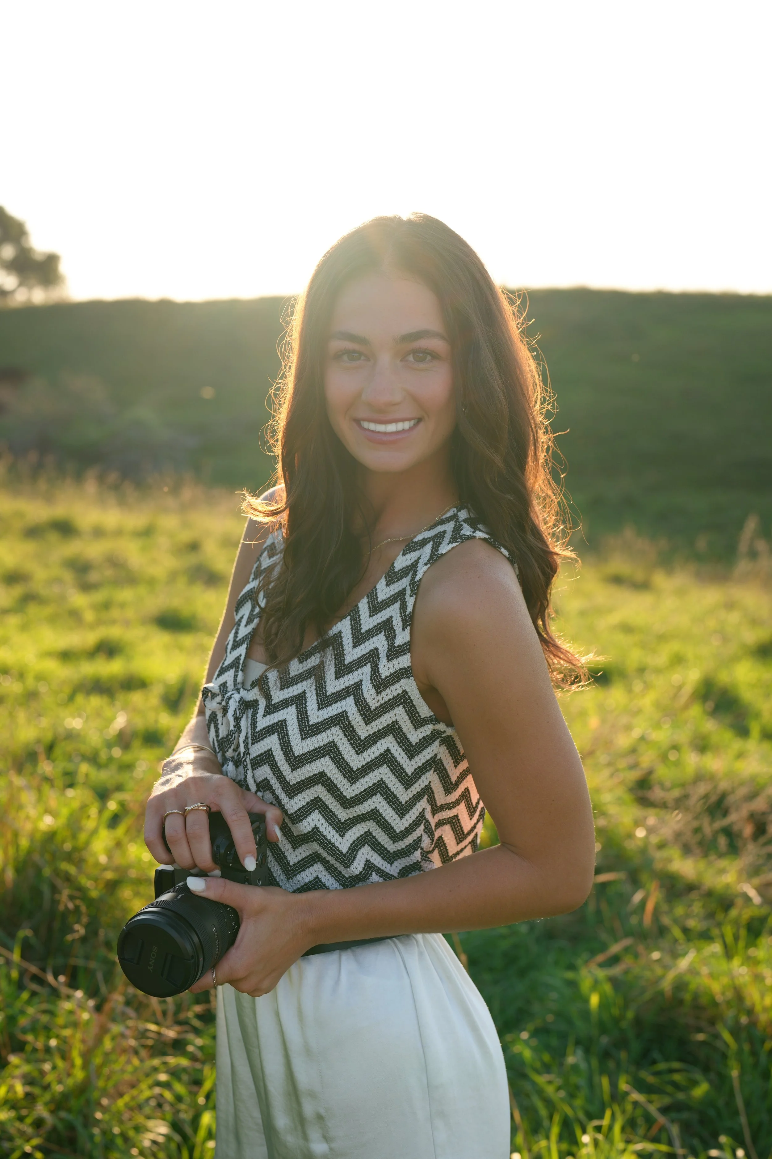 A young woman standing outdoors in a grassy field during sunset, holding a camera and smiling at the camera.