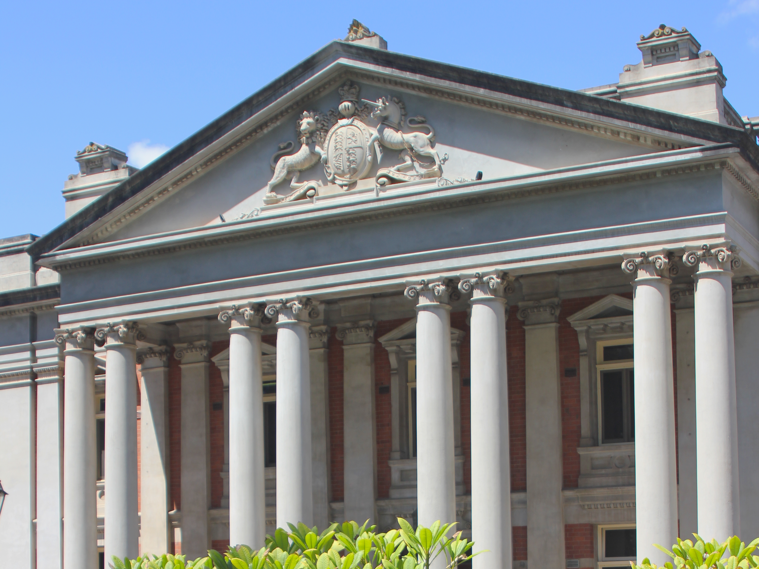 Front view of a historic building with classical columns, a detailed triangular pediment with emblem, two lions, and crown symbols, against a blue sky.