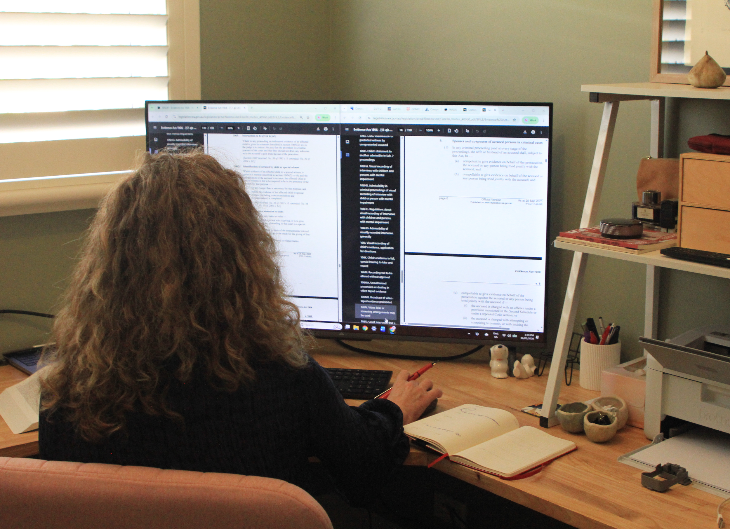 A woman with curly hair sitting at a desk with a dual monitor setup, reviewing legal documents with notes in an open notebook, in a well-lit room with blinds and organized office supplies.