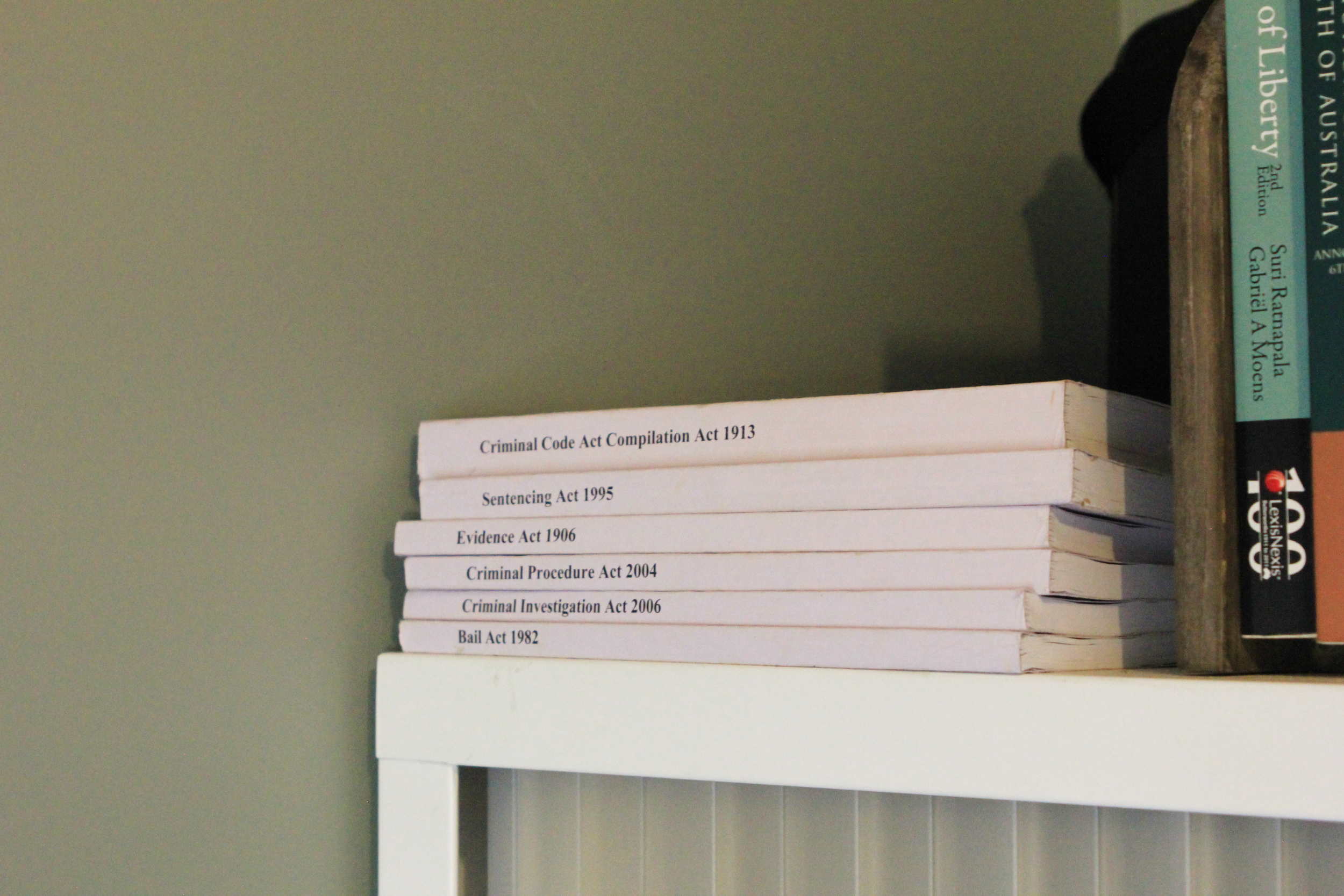 Stack of law books on a white shelf, with visible titles including 'Criminal Code Act Compilation Act 1913' and others related to criminal law, beside a wooden table with additional books.