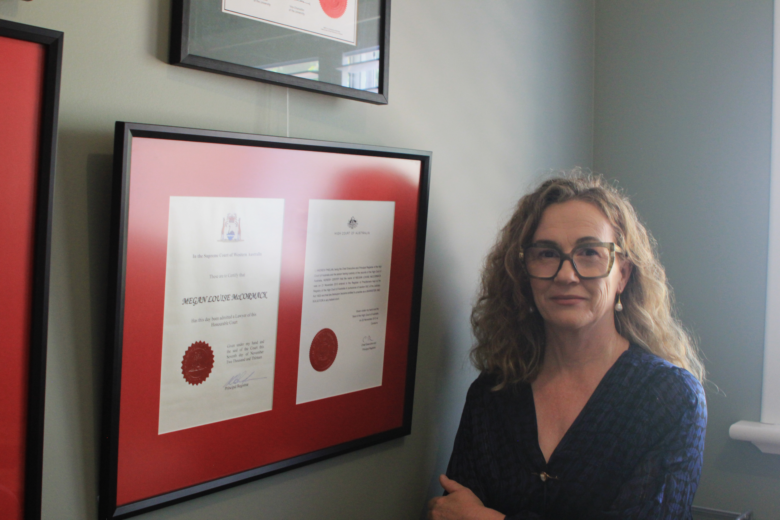 Woman with curly hair, glasses, and pearl earrings standing next to framed certificates on a wall.