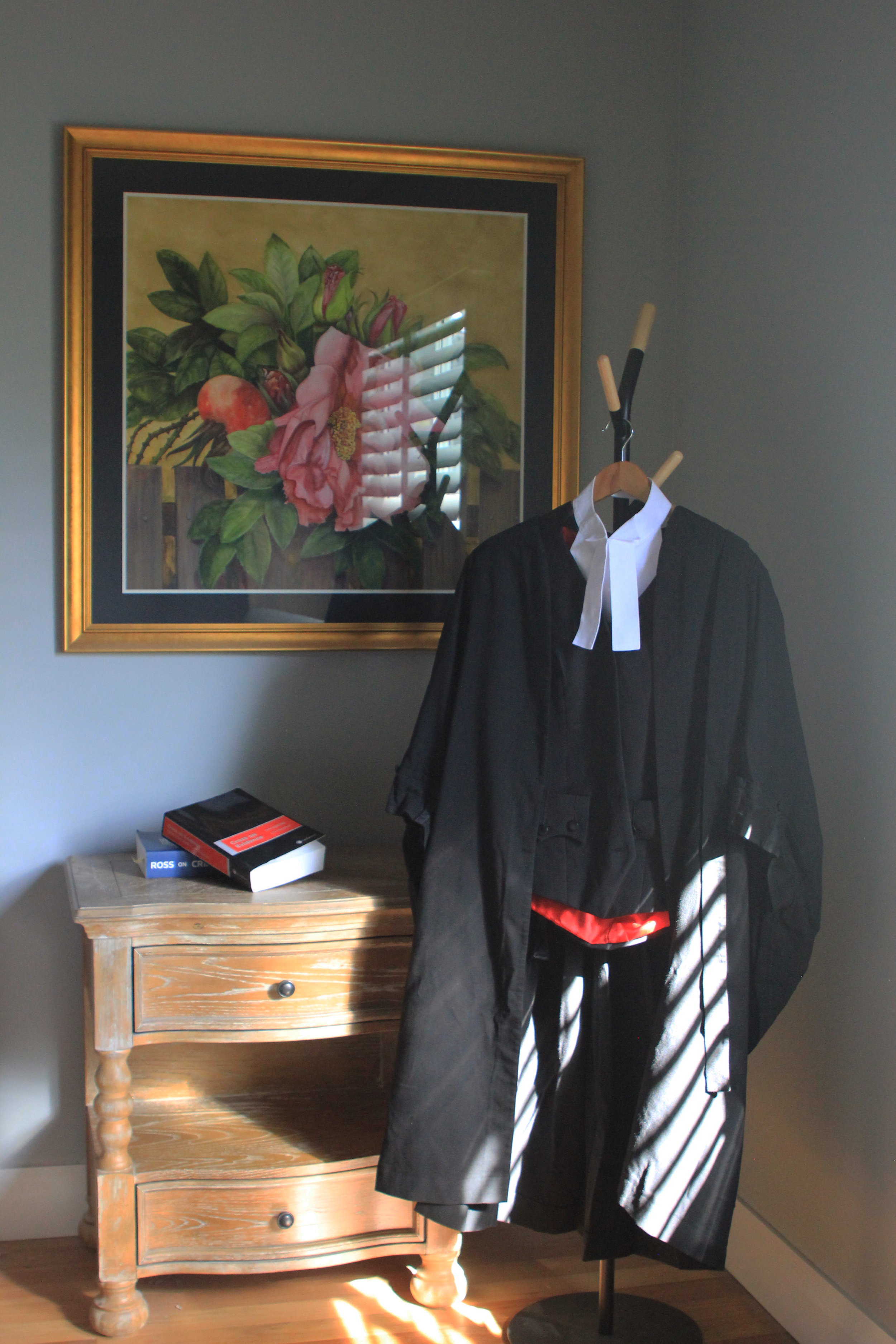A judge's black robe with a white collar hanging on a stand inside a room with a wooden desk, books, a painting of pink flowers on the wall, and sunlight streaming through window blinds.