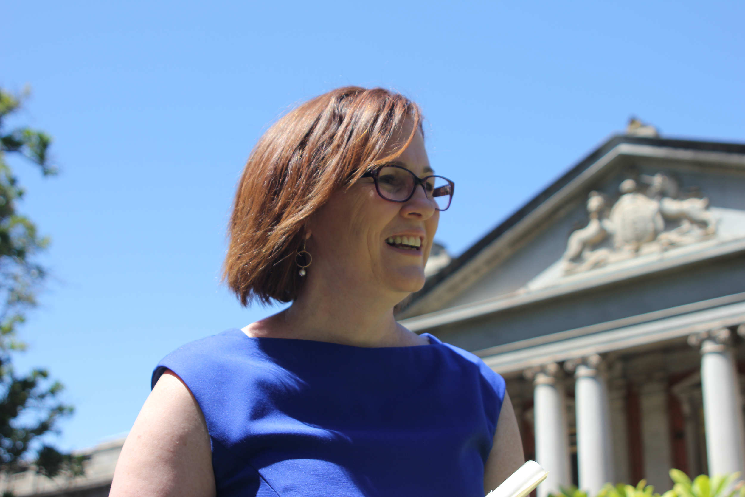 A photo of Megan McCormack in 2013, at her admission into legal practice. She is wearing a blue dress, with glasses and shoulder length auburn hair. She is in front of the Supreme Court of Western Australia; the sky is bright blue.