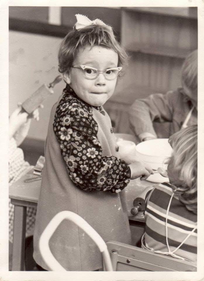 A black and white photo of Megan McCormack in 1973, aged approximately 4 years, wearing glasses, a bow in her hair standing at a table with other children. She is holding a bowl.