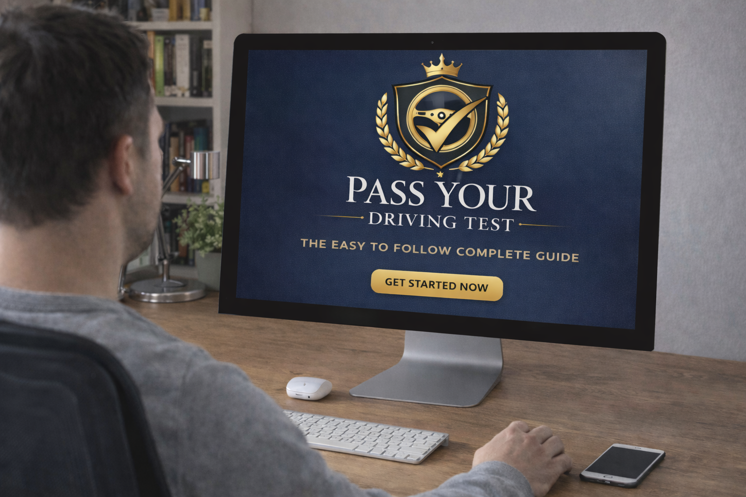 Man sitting at a wooden desk using a computer to access a driving test website titled 'Pass Your Driving Test.' The screen displays a shield with a checkmark, a crown, and the text 'Get Started Now.'