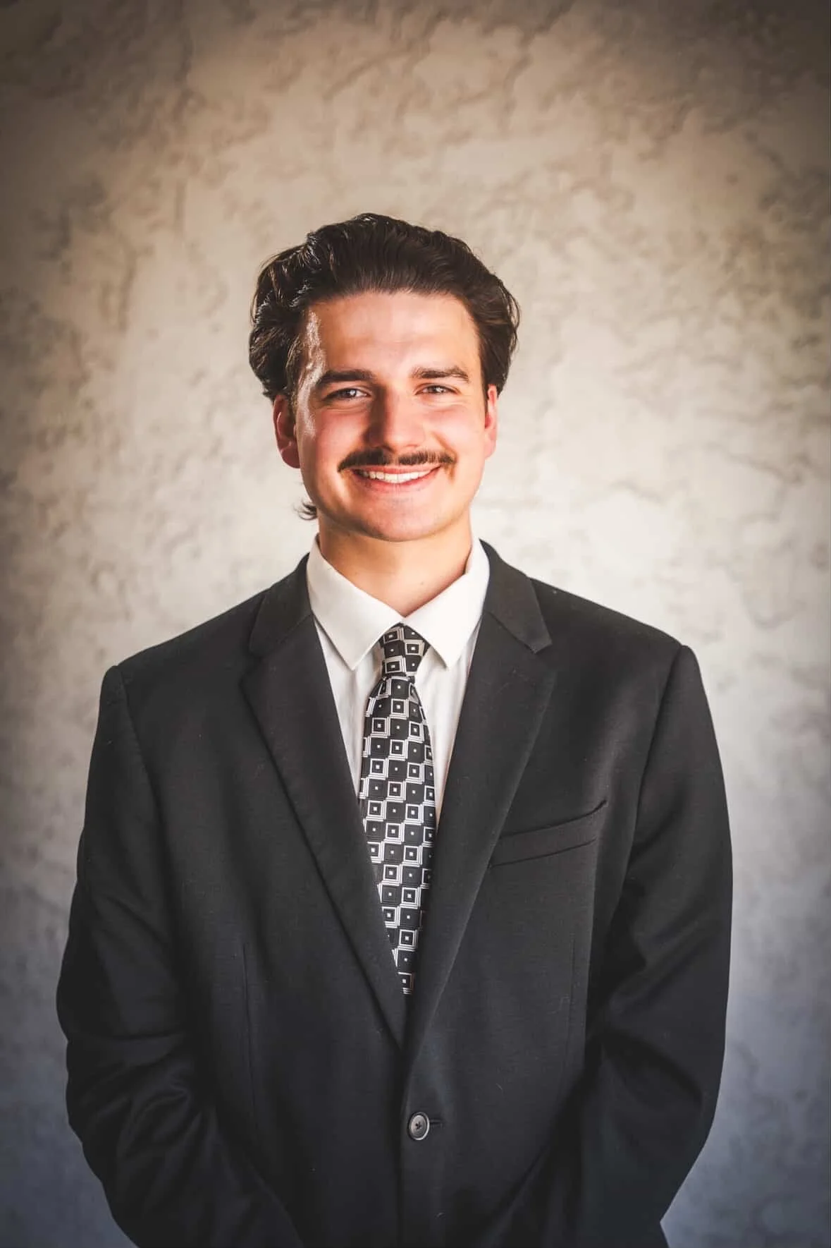 A young man dressed in a black suit with a patterned tie and white shirt, smiling at the camera, standing against a textured, light-colored background.