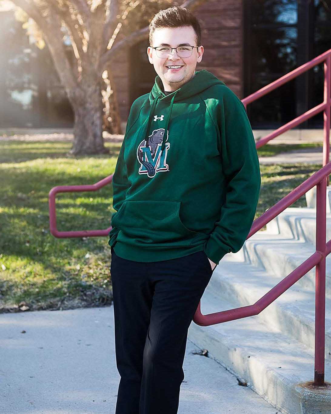 A young man with glasses smiling, standing outdoors with sunlight behind him, wearing a green hoodie with a sports team logo, dark pants, and leaning against a pink railing.