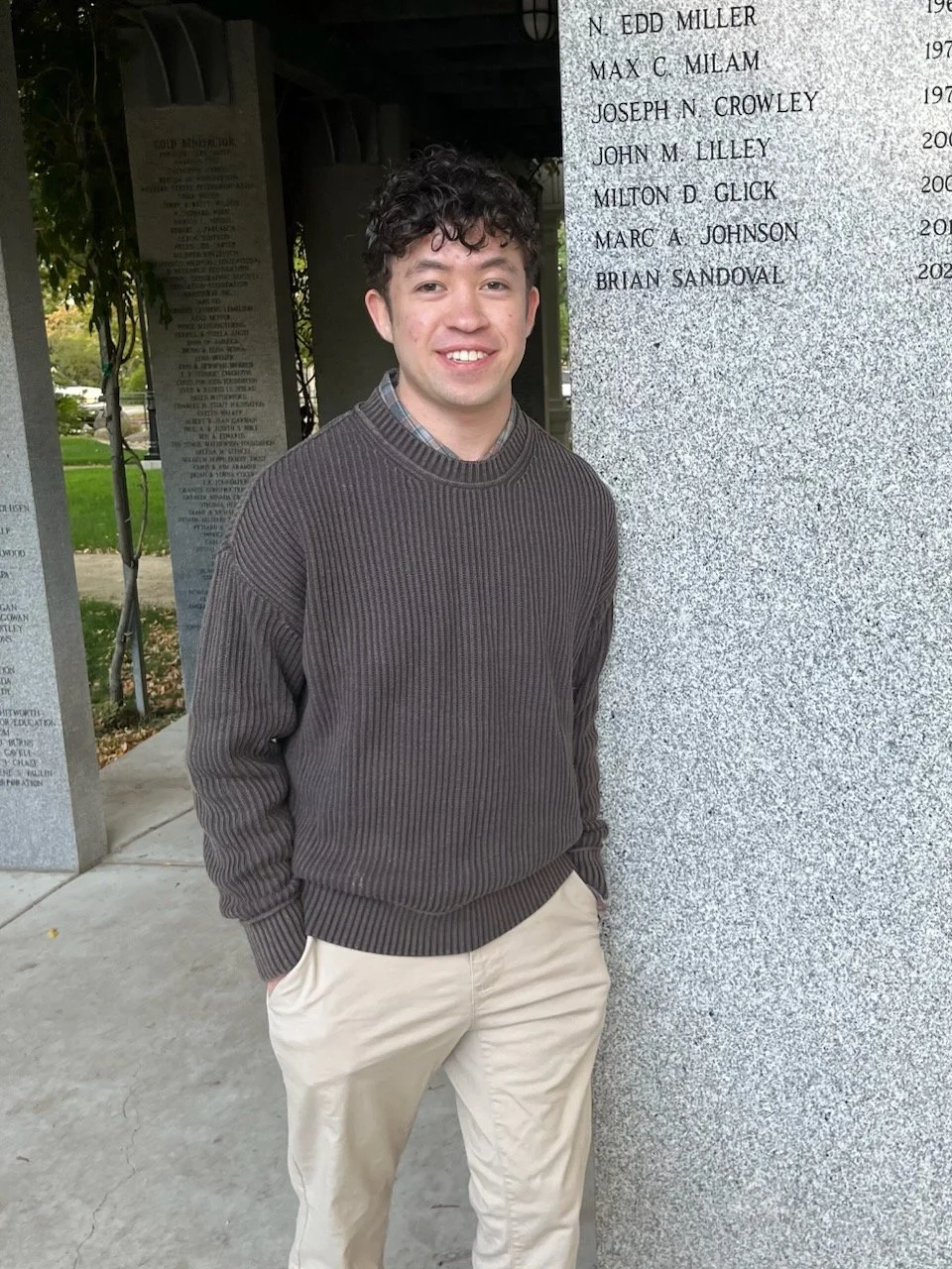 Young man with curly dark hair, dressed in a brown ribbed sweater and khaki pants, smiling and standing beside a gray memorial wall with names inscribed.