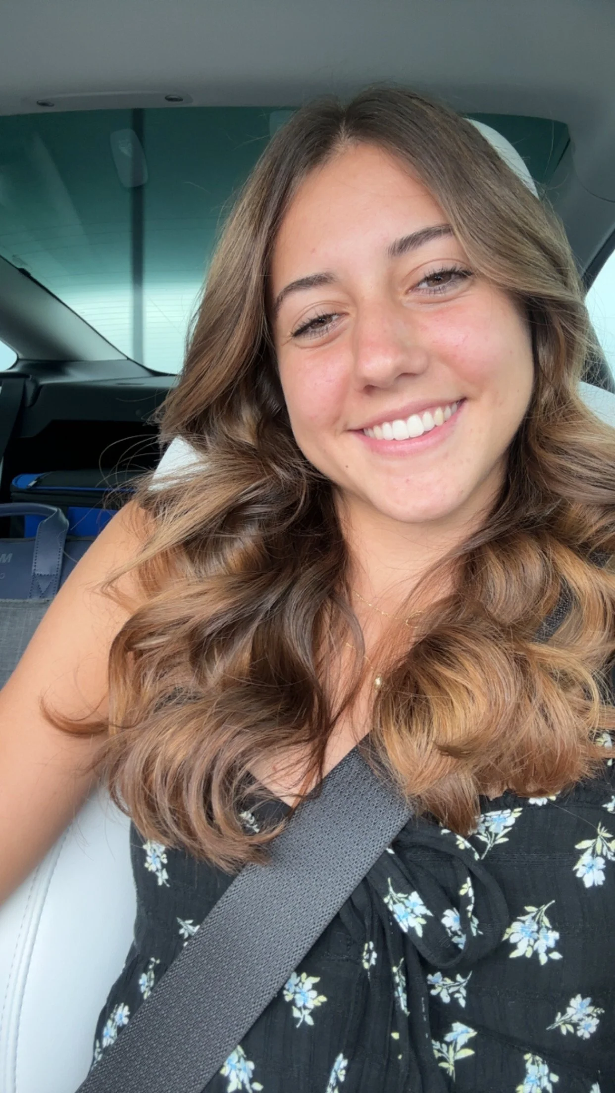Young woman with long, wavy brown hair smiling in a car, wearing a black floral dress and a seatbelt.