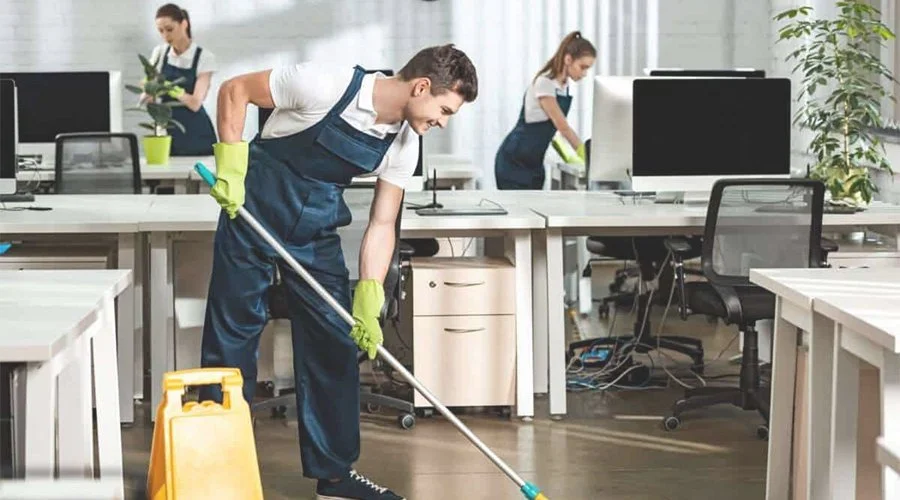 Man in a blue apron and yellow gloves cleaning the office floor with a mop in an office setting. Two women are working at their desks in the background.