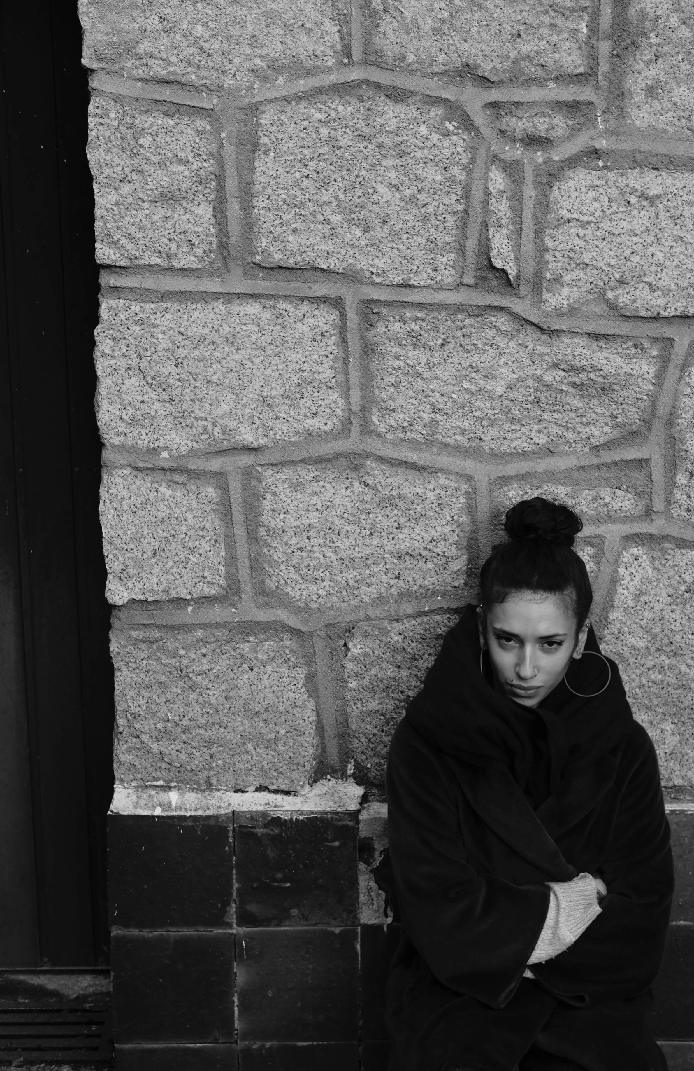 A young woman with dark hair styled in a bun, wearing a dark hoodie and light-colored gloves, sitting against a stone wall with a serious expression.