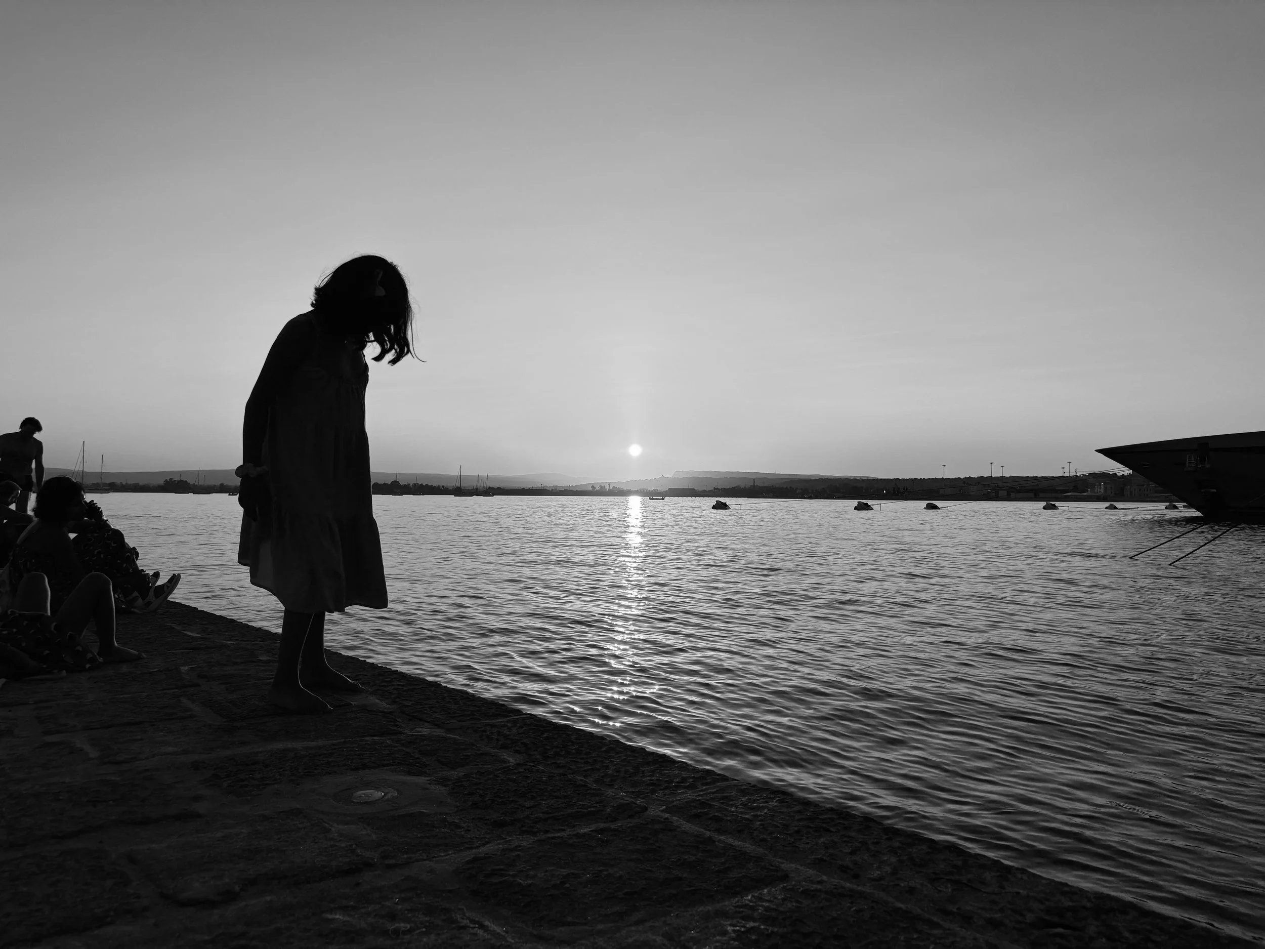 Silhouette of a woman standing by the water at sunset with boats in the distance.