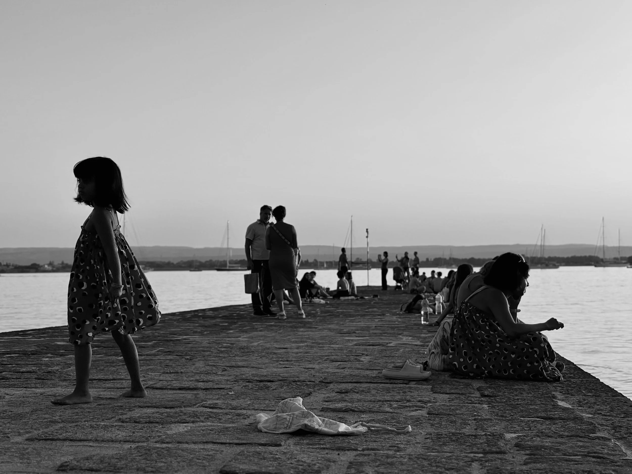 A black and white photo of people by a body of water, with some sitting and others standing. A girl in a polka-dot dress walks barefoot on the ground in the foreground, while others are gathered or walking near the water, with sailboats and a distant