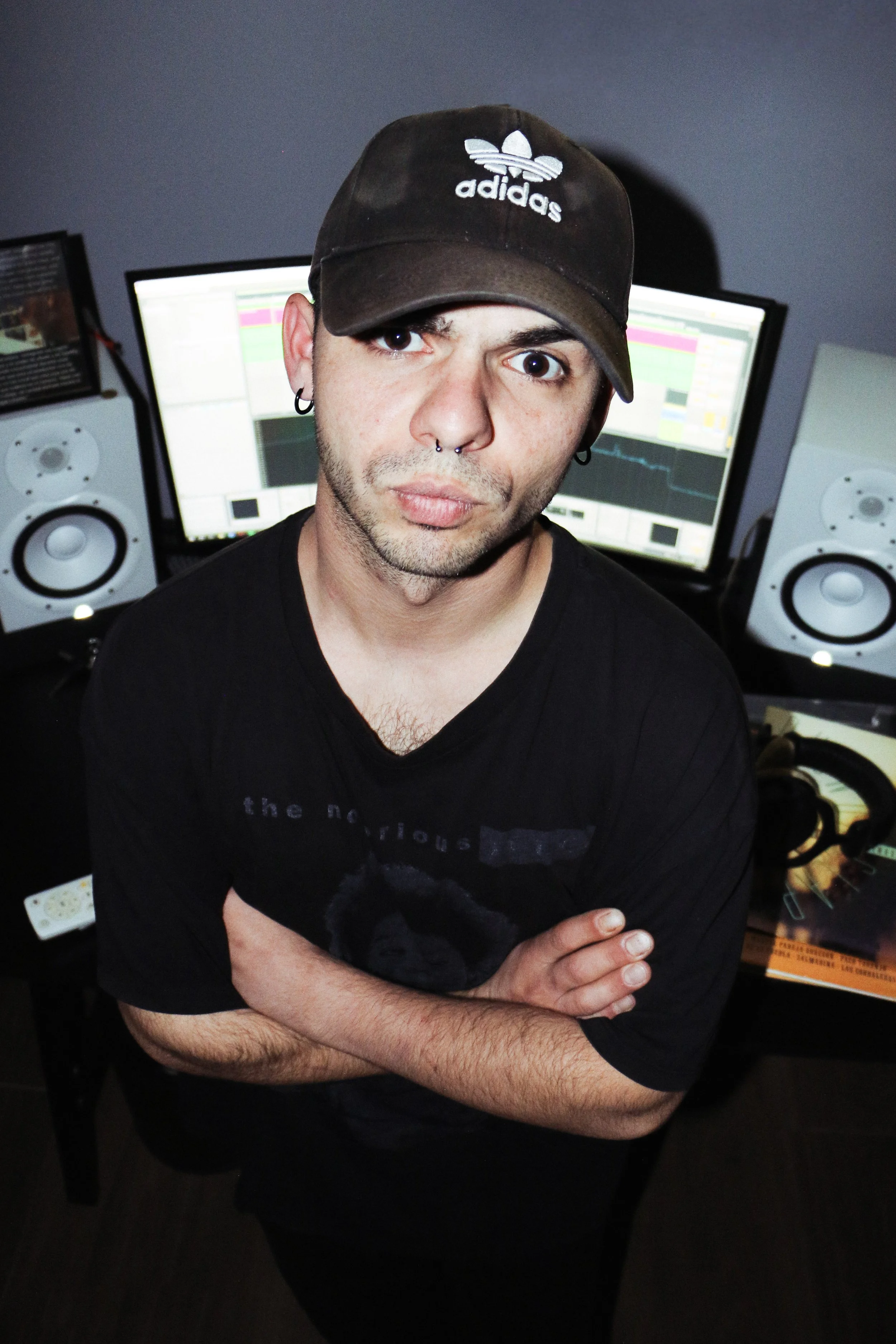 A young man with dark hair, wearing a black t-shirt and a brown Adidas cap, stands with his arms crossed in front of two computer monitors with colorful waveforms, in a dimly lit room with music speakers.