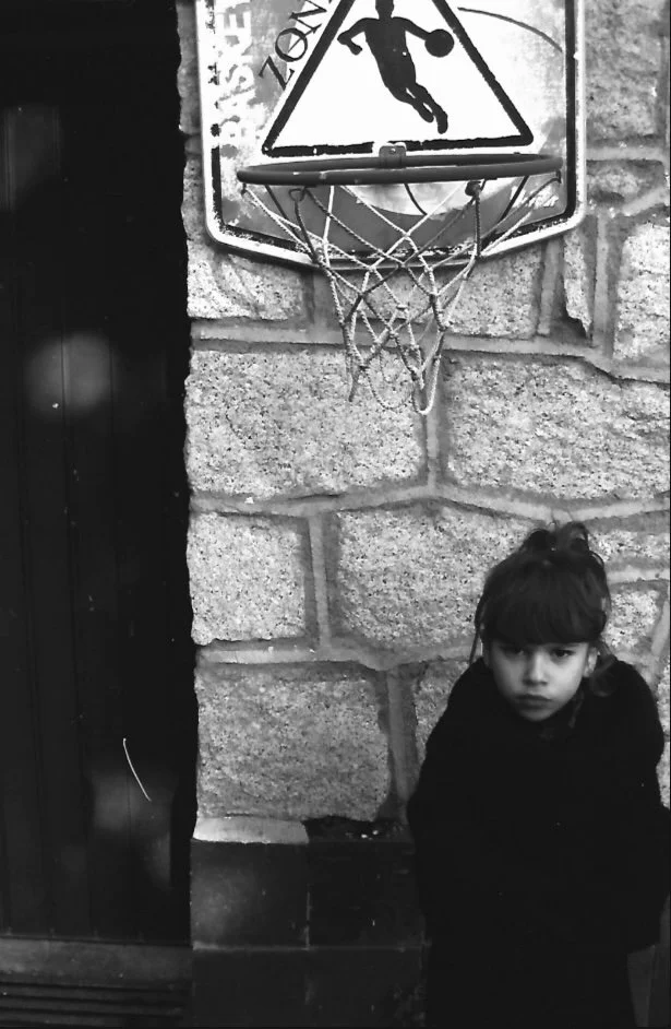 A young girl with dark hair and a serious expression, standing in front of a stone wall, next to a basketball hoop with a worn net and a sign above it showing a silhouette of a person running.