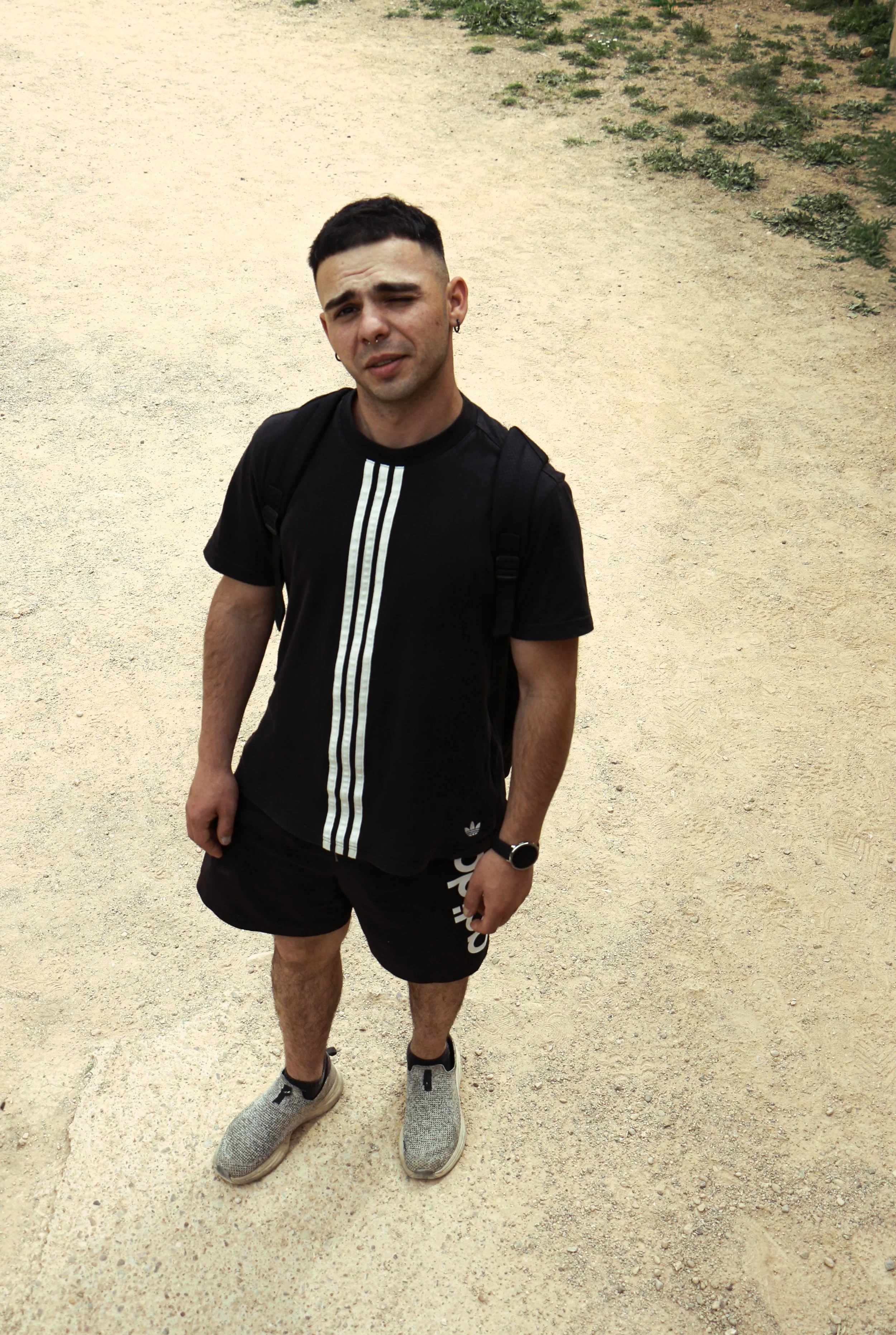 A young man with short dark hair, light skin, and a septum piercing stands outdoors on a dirt pathway with some green plants on the side. He is wearing a black Adidas t-shirt, black shorts, gray sneakers, a wristwatch, and a backpack, and is making a