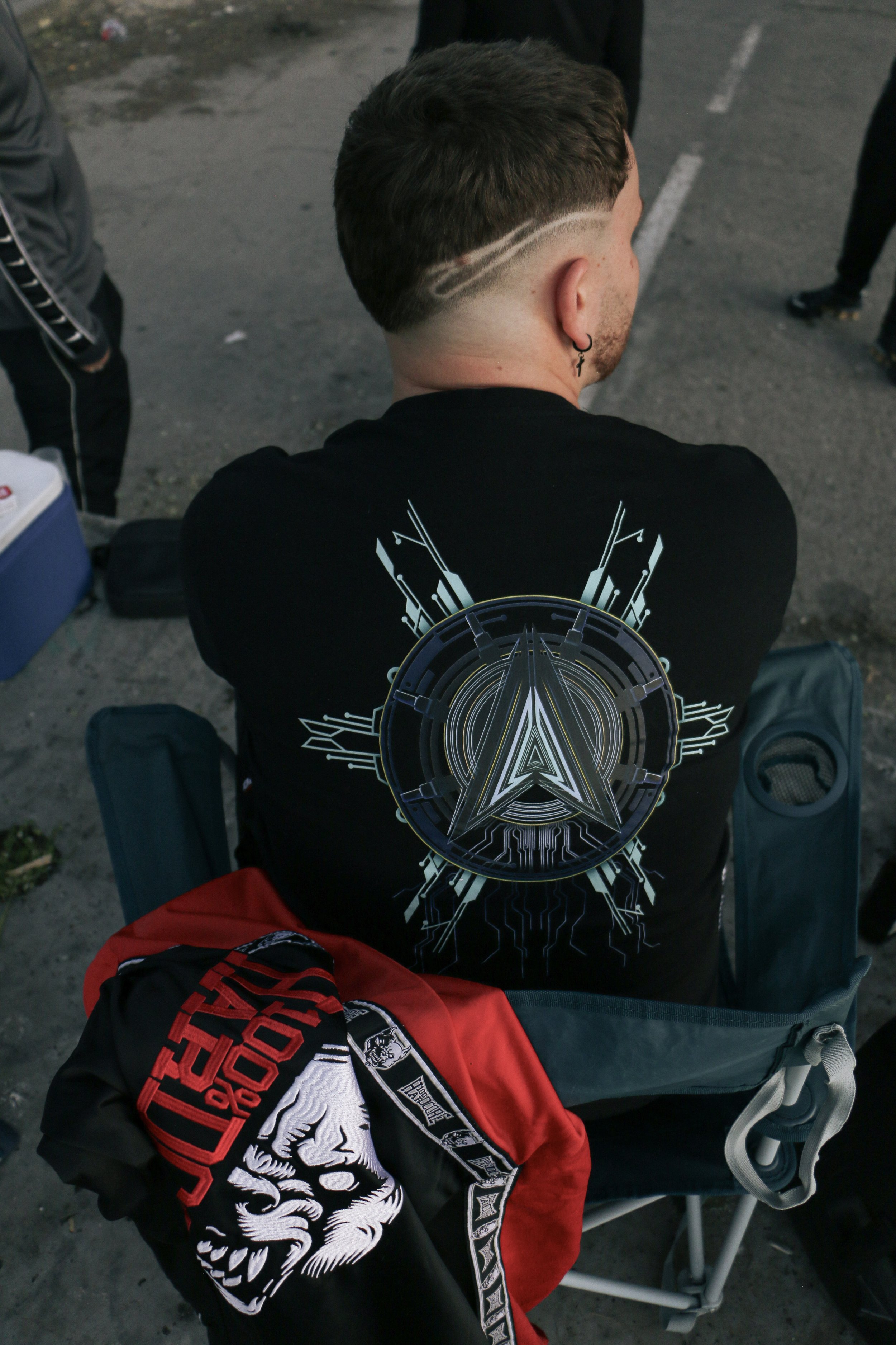 A person with short brown hair, wearing a black T-shirt with a large circular graphic and a white skull and eagle design on the back, is seated outdoors on a gray street or parking lot. The person has two parallel lines shaved into the side of their 