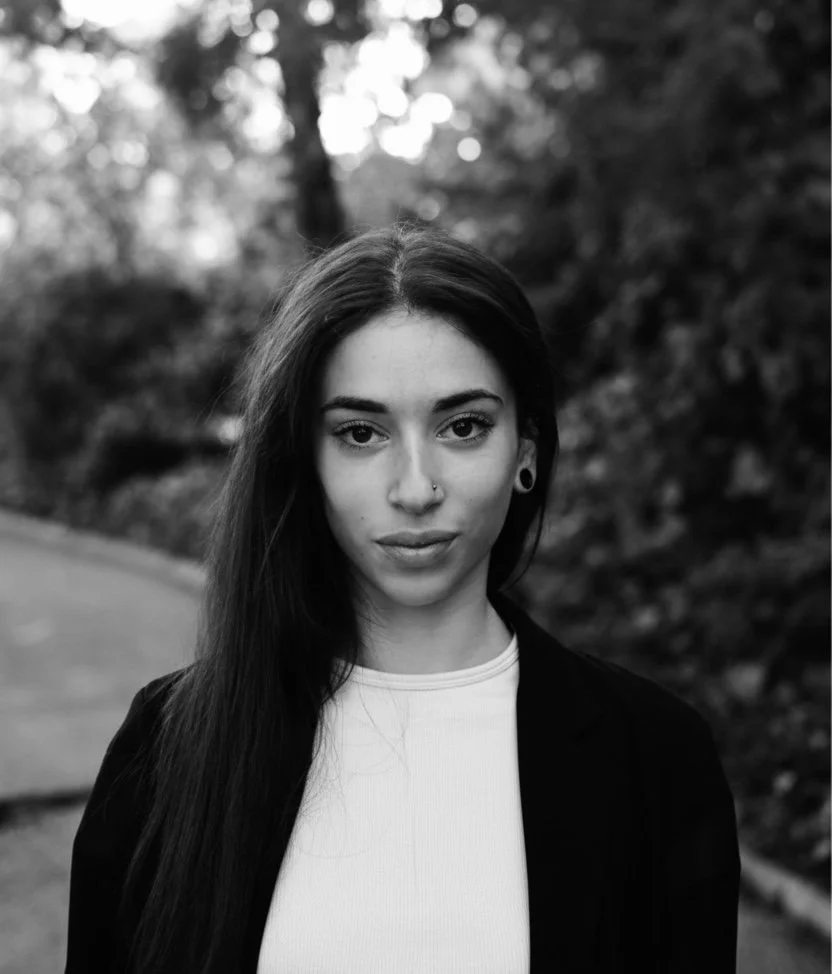 Black and white portrait of a young woman with long dark hair, wearing a nose piercing and earrings, standing outdoors with blurred trees in the background.