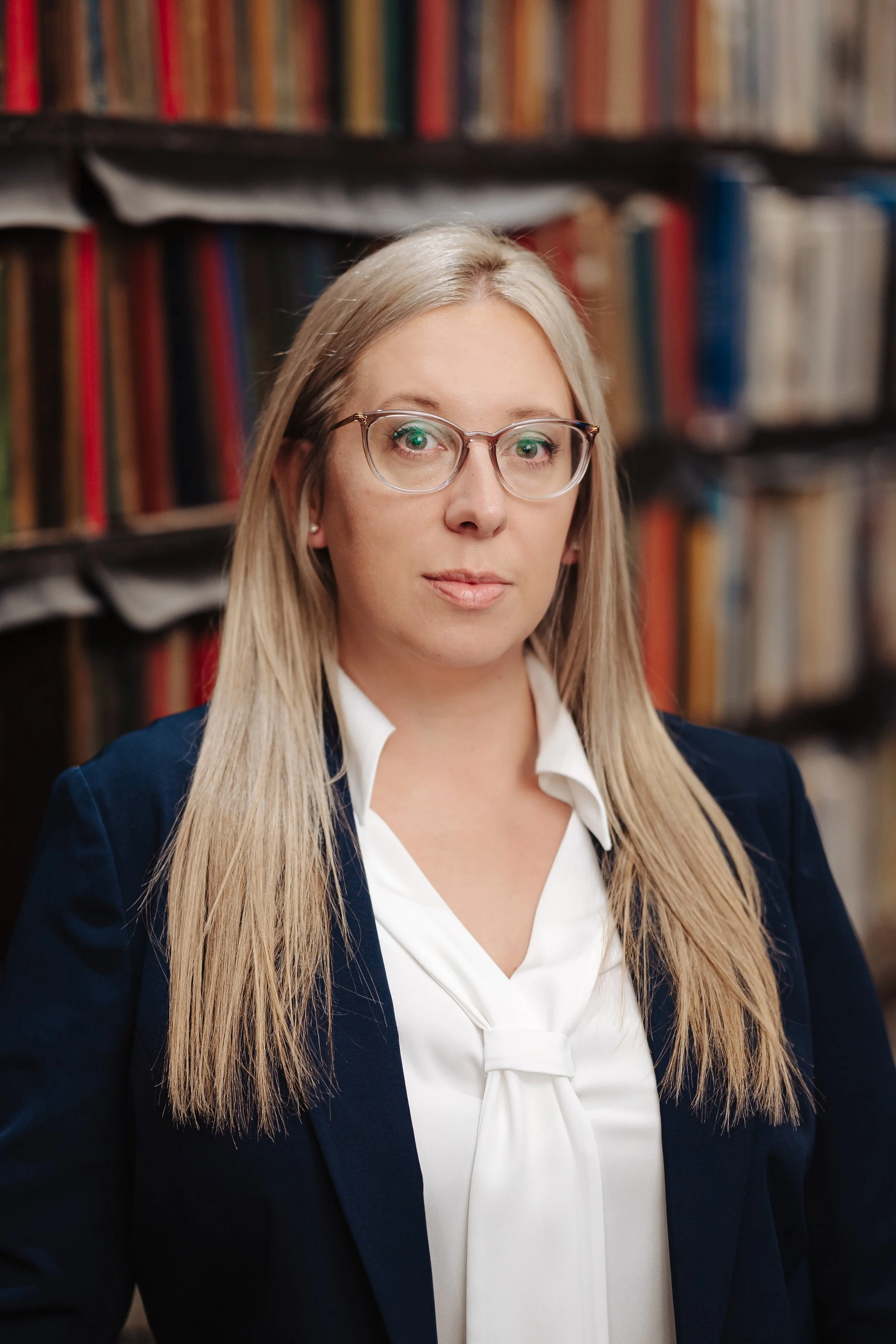 Dr Jessica Taylor - A professional woman with long blonde hair, glasses, and a white blouse, standing in front of a bookshelf filled with books.