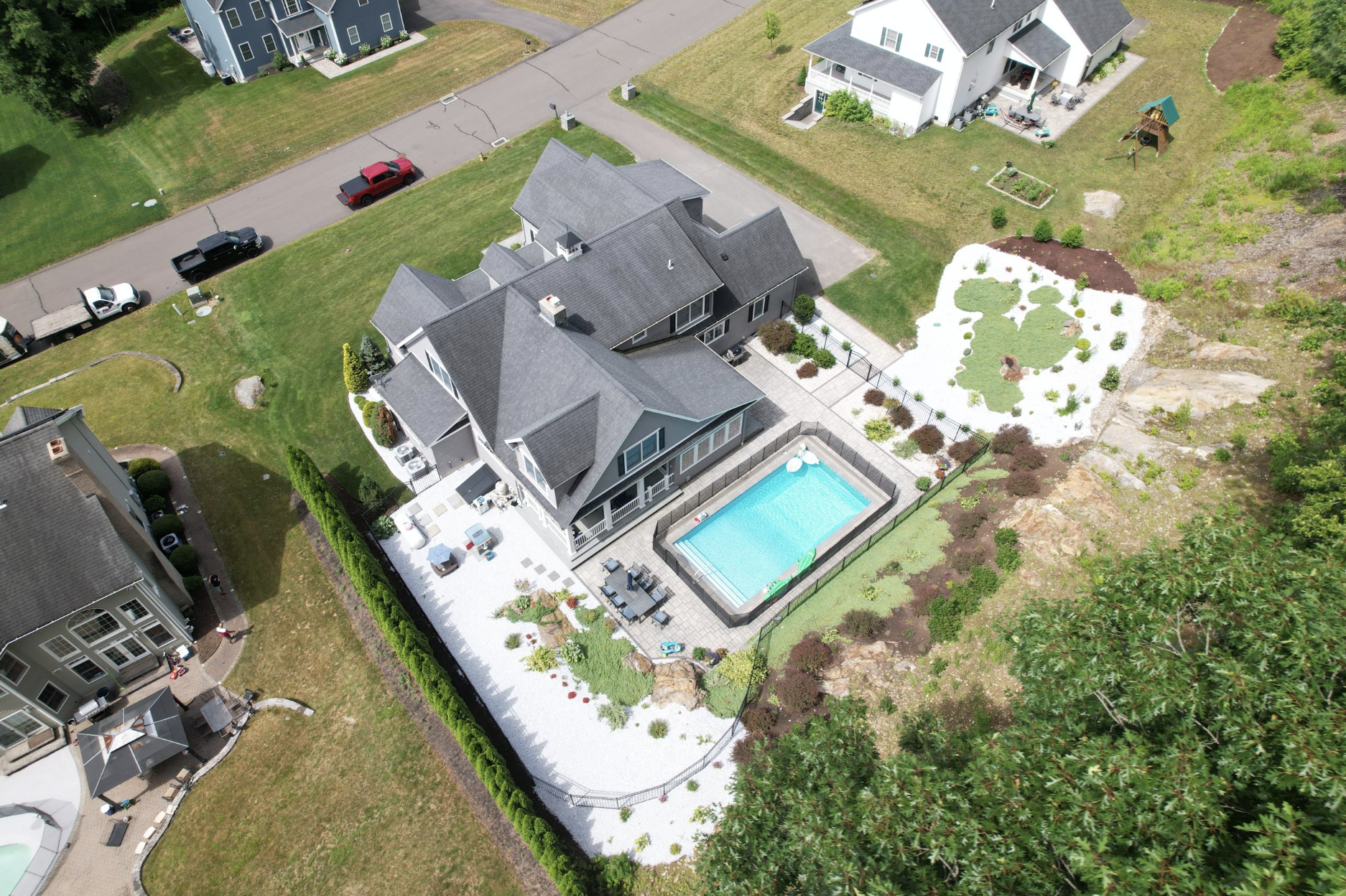 Aerial view of a suburban backyard with a swimming pool, a patio area with outdoor furniture, a landscaped garden, and a neighboring house.