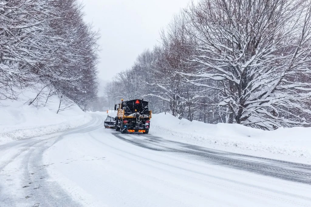 Snowplow clearing snow from a curved road lined with snow-covered trees on a winter day.
