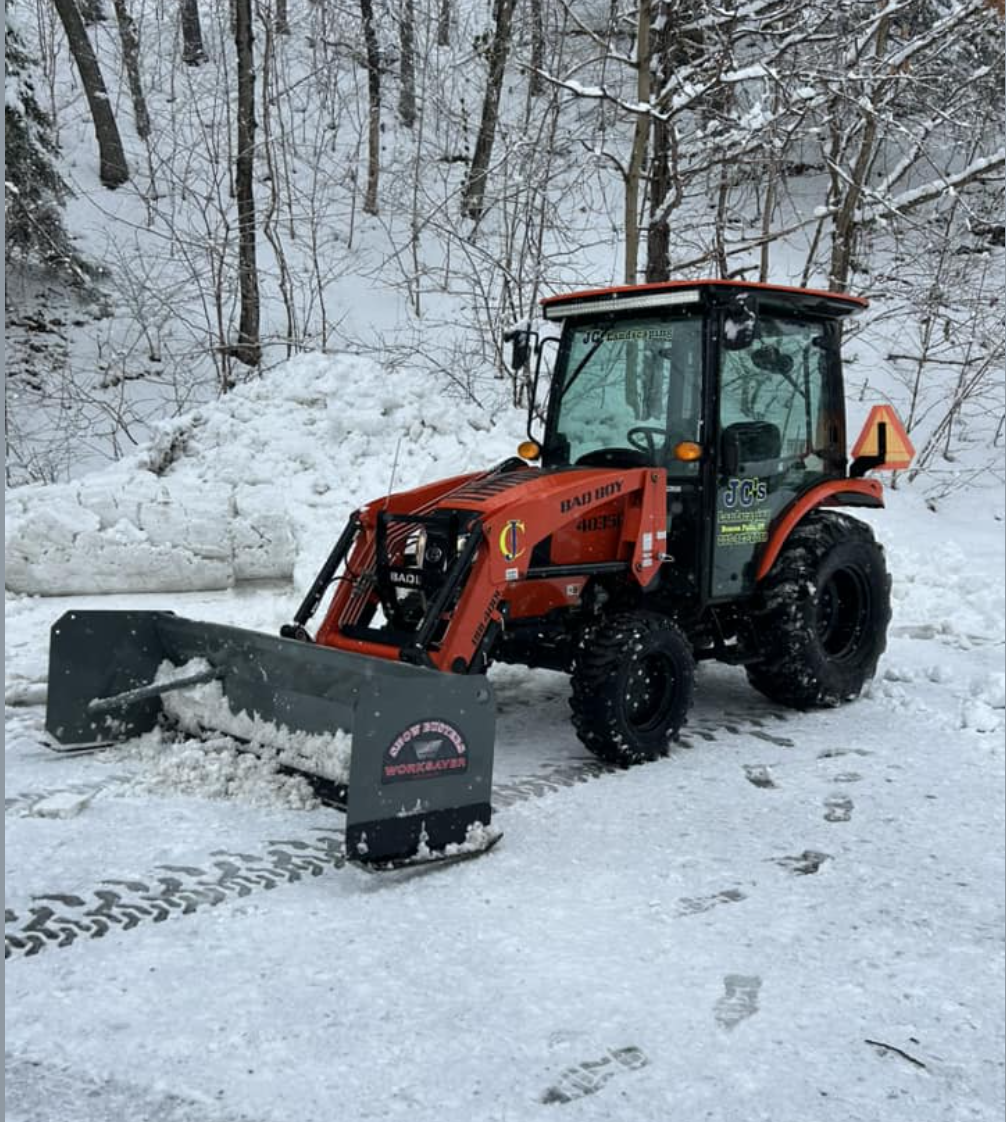 A red tractor equipped with a snow plow blade clearing snow from a road in a snowy wooded area.