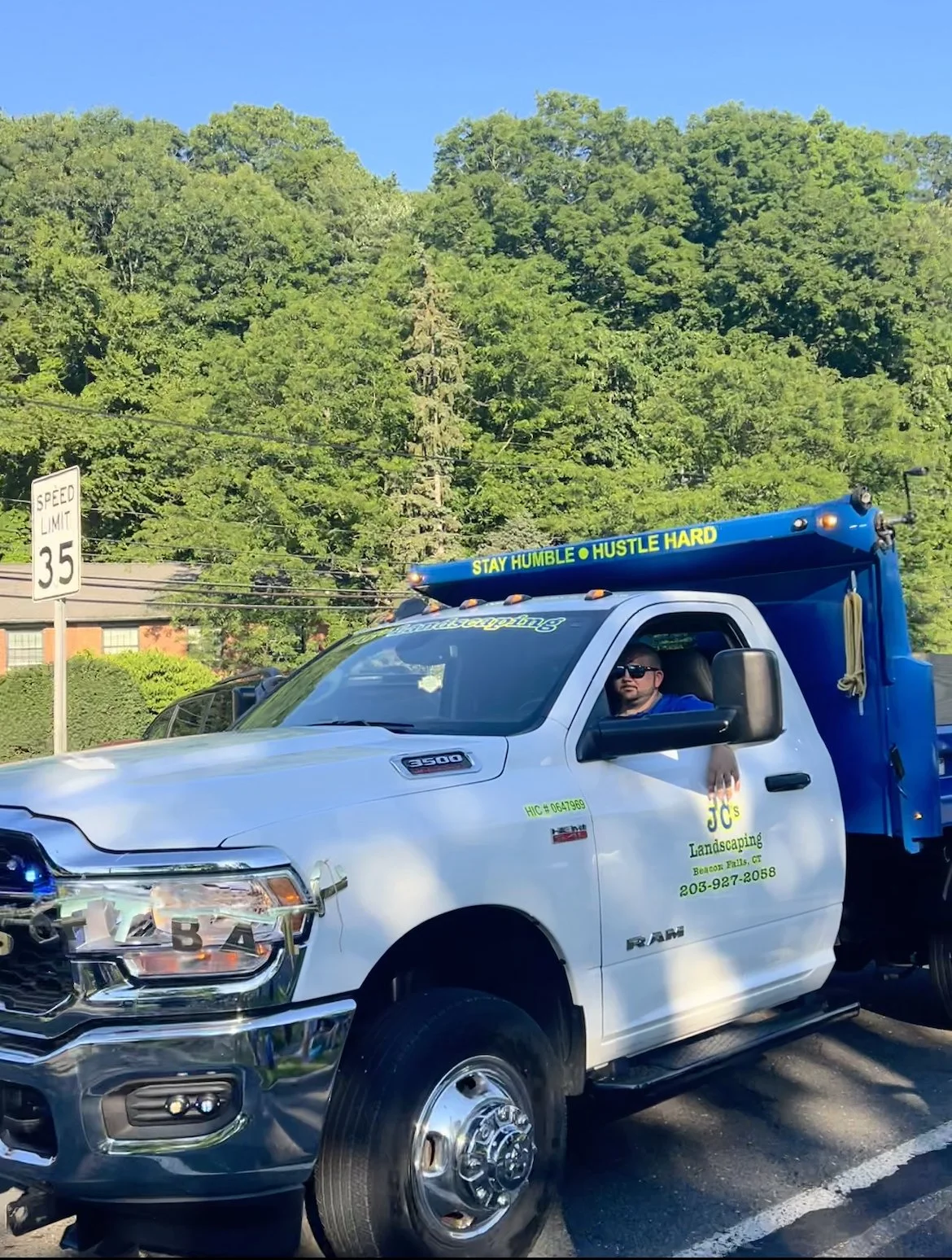 A white RAM pickup truck with landscaping company branding parked on a street. The truck has a blue utility box on the back, with a sticker that reads 'STAY HUMBLE • HUSTLE HARD.' A man wearing sunglasses is sitting in the driver's seat. There is a speed limit sign of 35 mph and a green hillside with trees in the background.