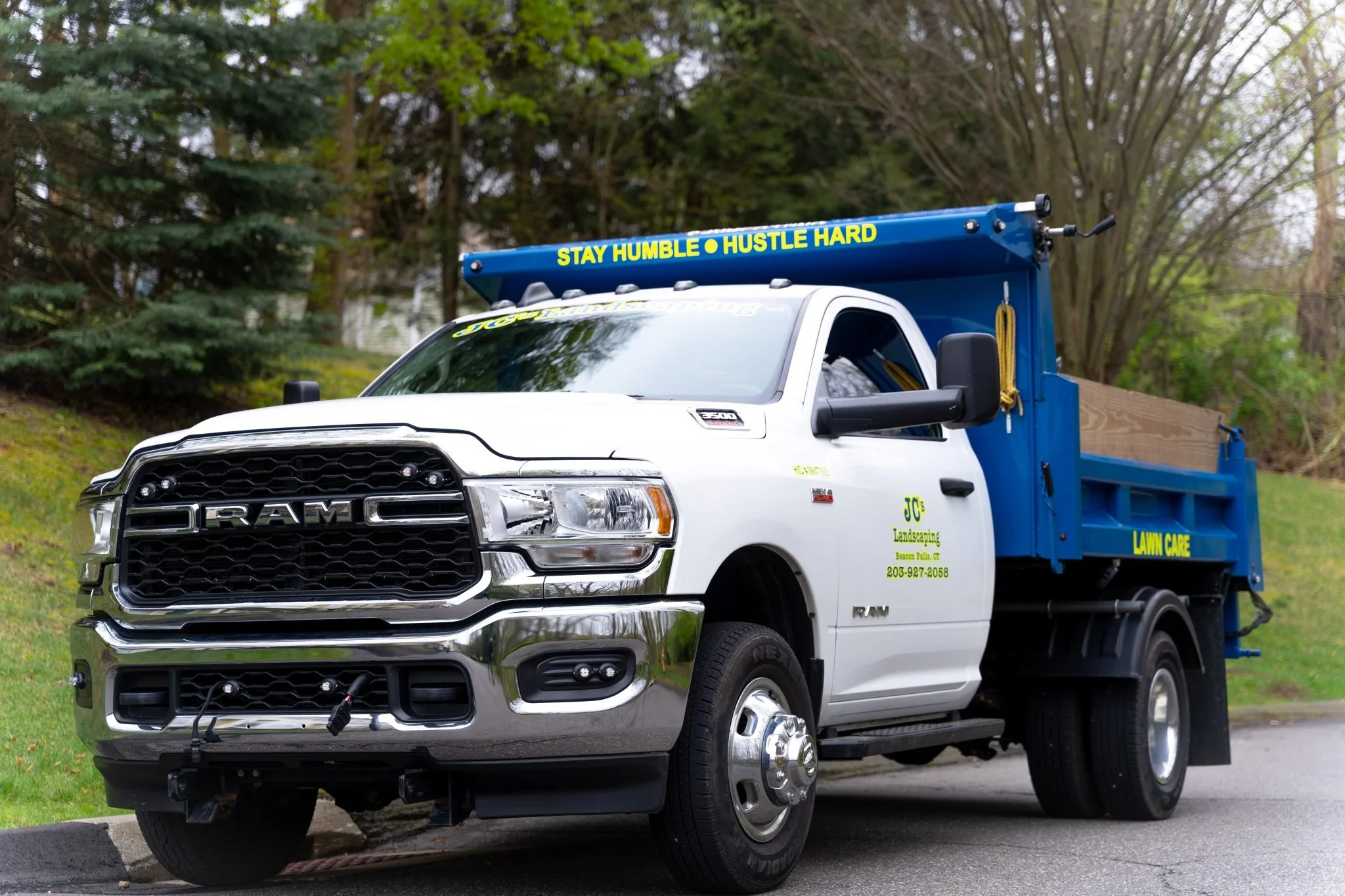 A white RAM pickup truck with a blue flatbed and yellow text, parked on a driveway in front of green trees. The truck has a sign that reads 'STAY HUMBLE ● HUSTLE HARD' on the top edge of the flatbed and 'LAWN CARE' on the side. The side of the truck displays contact information for J.C's Landscaping.
