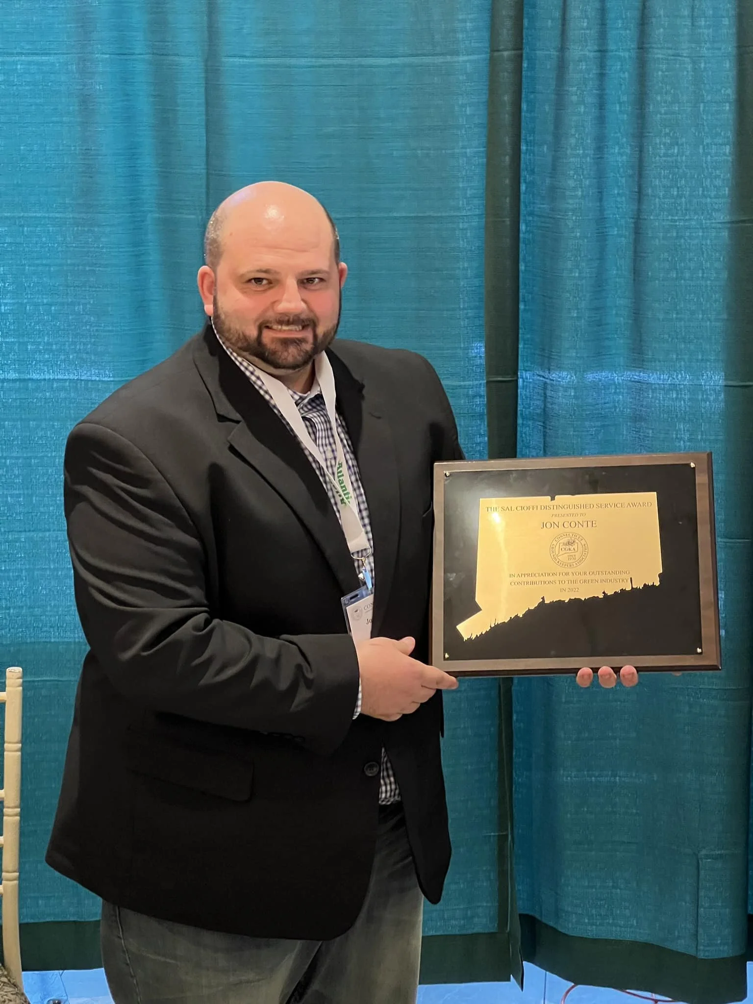 A man in a dark suit and checkered shirt holding an award plaque with a yellow and black design.
