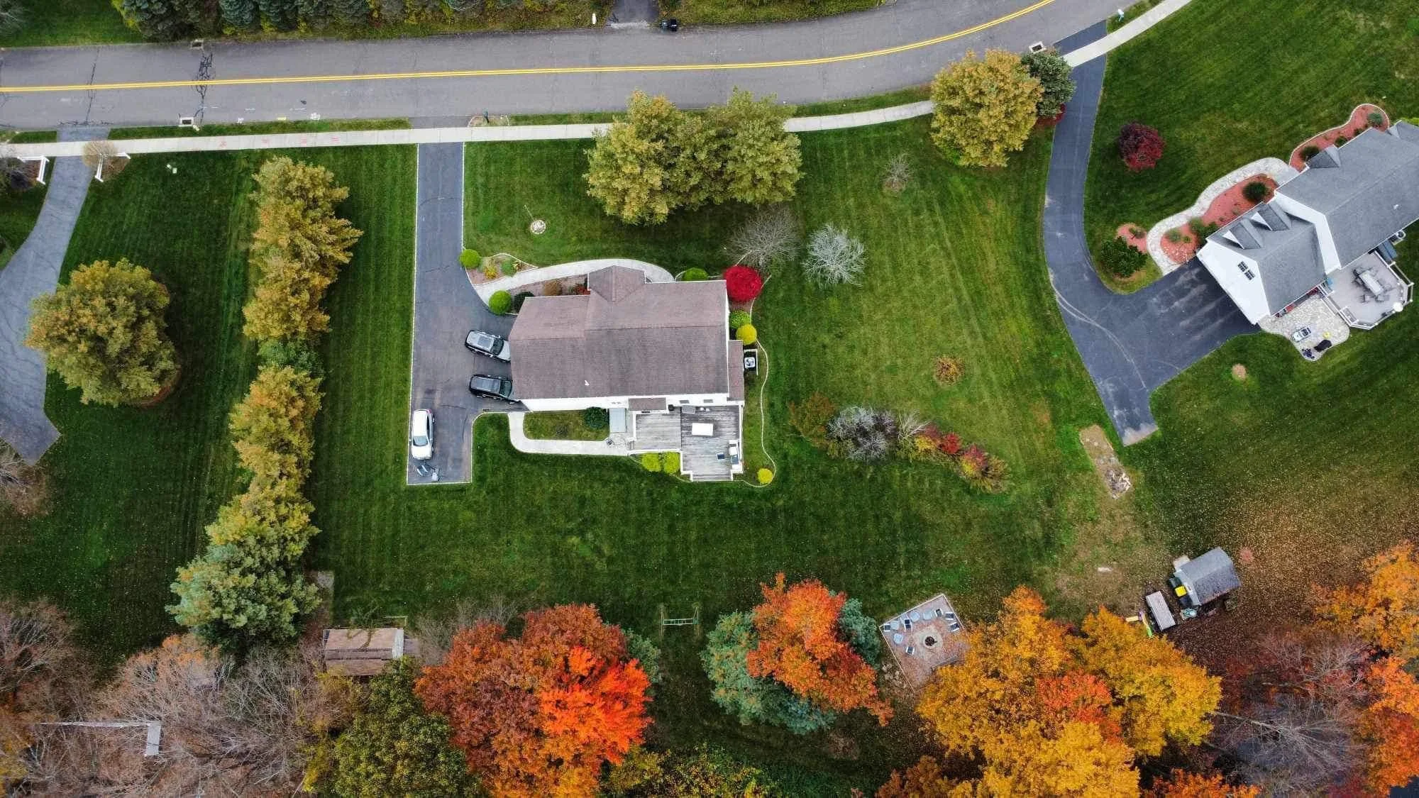 An aerial view of a residential neighborhood during fall, showing a house with a driveway, several cars, a lawn, and trees with colorful autumn leaves.