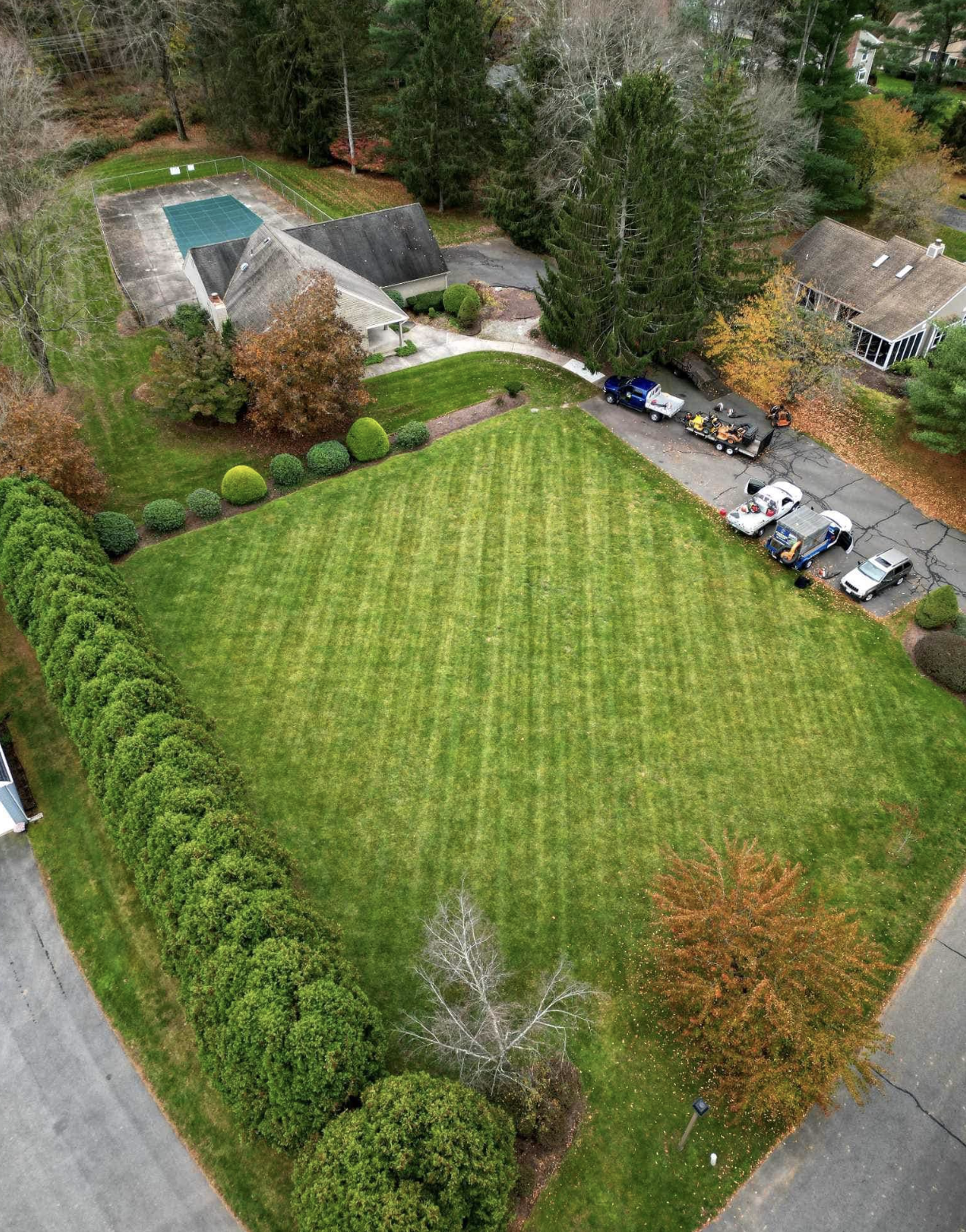 Aerial view of a large, well-maintained green lawn surrounded by trees and shrubs. There is a paved parking area with several parked vehicles and a trailer. A small building with a pitched roof is visible, along with a fenced tennis or basketball court. Some trees show fall foliage.