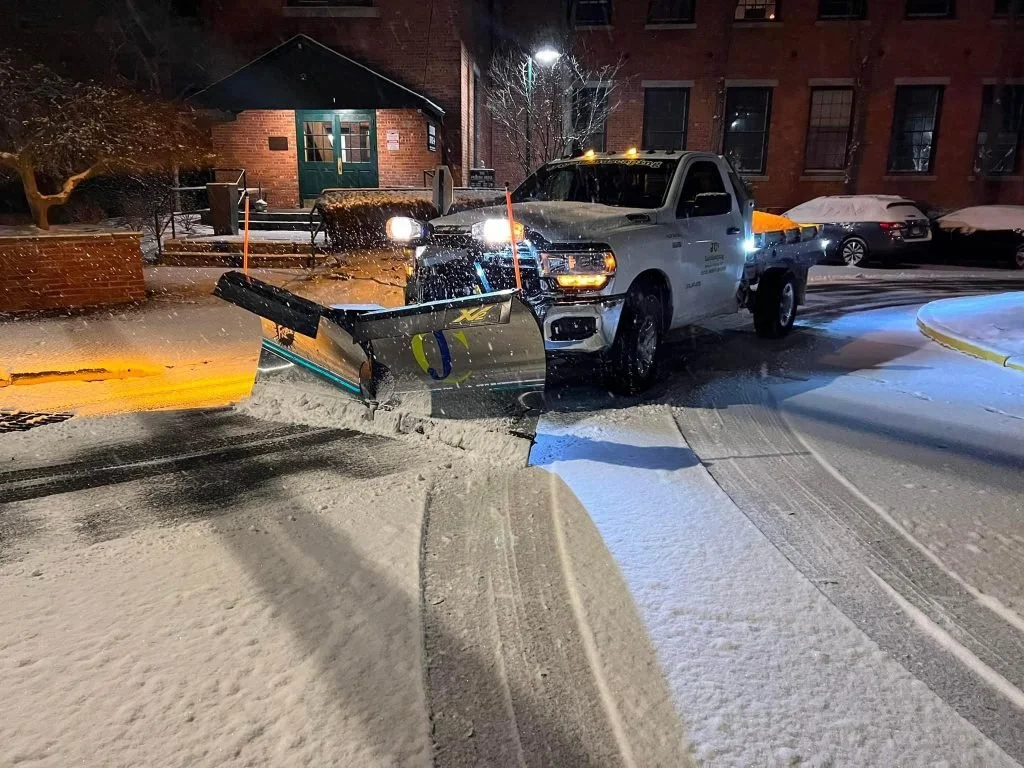 A pickup truck equipped with a snow plow attachment clearing snow from a city street at night. Snow is falling and ground is covered in snow. Residential buildings and parked cars are in the background.