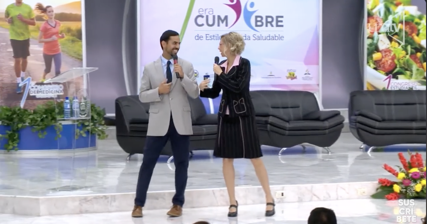 A man in a light gray suit and a woman in a striped black skirt and blazer are speaking into microphones on stage, smiling at each other. There are black chairs, a flower arrangement, and banners with images and text in the background.