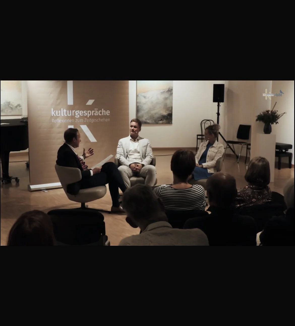 Drei Personen im Gespräch bei einer Diskussionsrunde im Raum, im Hintergrund eine Wand mit Gemälden und ein Banner mit der Aufschrift 'kulturgespräche'.