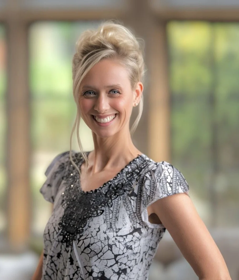 Eine lachende blonde Frau mit hochgestecktem Haar in einem Raum mit großen Fenstern und grüner Natur im Hintergrund, trägt ein schwarz-weiß gemustertes Kleid.