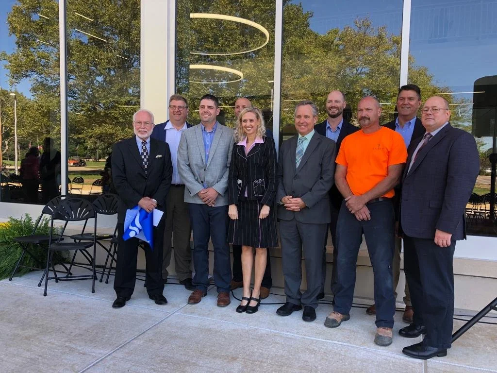 Eine Gruppe von zehn Menschen, bestehend aus Männern und einer Frau, posiert vor einem großen Fenster bei einer Veranstaltung. Sie sind in formeller und legerer Kleidung, und es ist Herbst oder Frühling, da die Bäume grün sind.