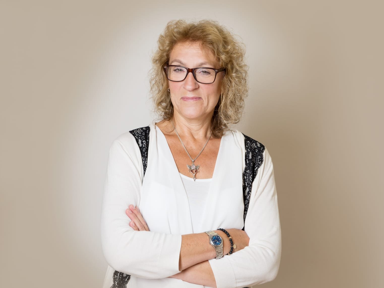 Ein älterer Frau mit lockigem, blondem Haar, Brille, weißem Top und schwarzem Schulterträger, die ihre Arme verschränkt hat, vor einem neutralen Hintergrund.